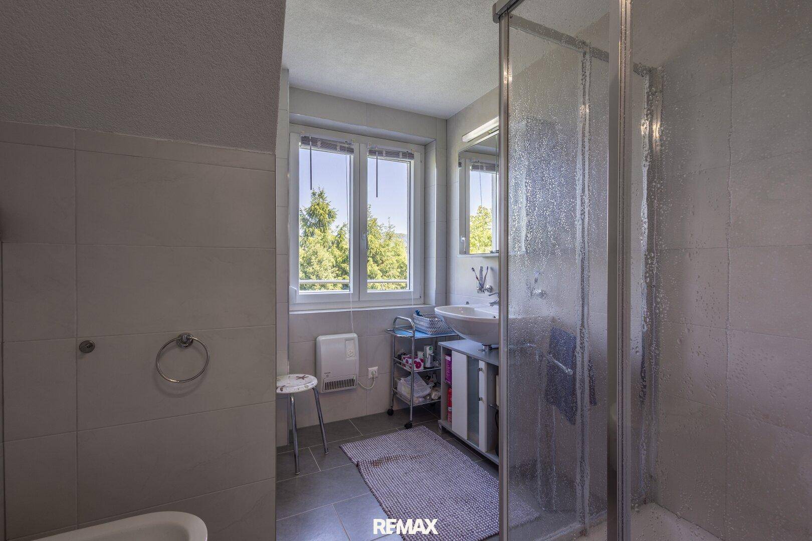 Badezimmer mit Dusche, Waschbecken und Fenster mit Blick ins Grüne.