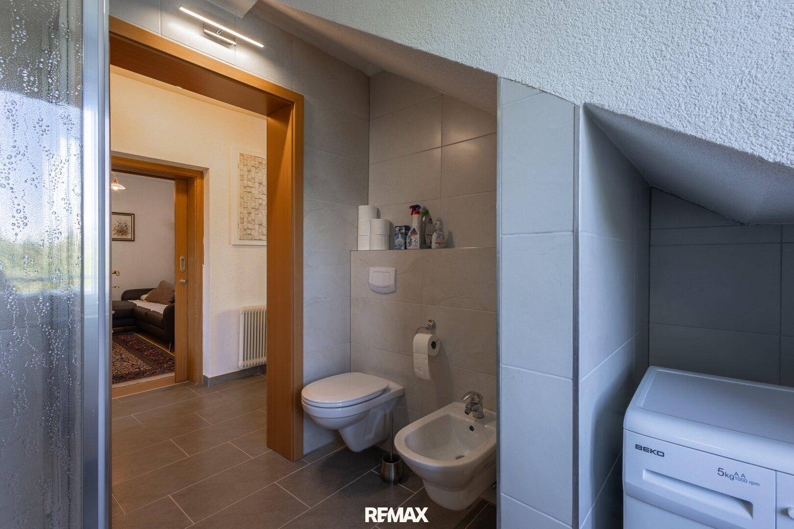 Badezimmer mit WC, Bidet und Waschmaschine, angrenzend an einen weiteren Raum.