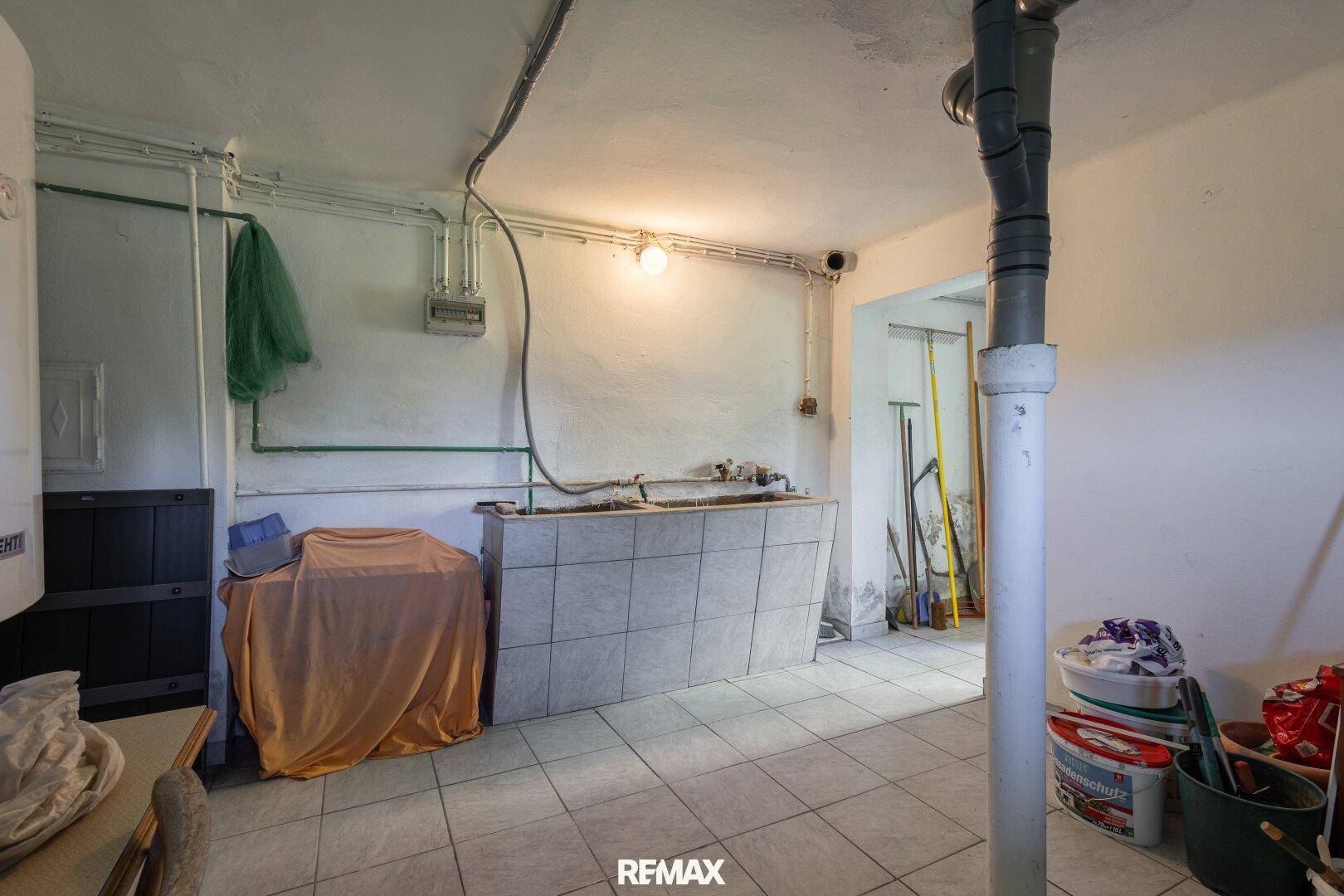 Kellerraum mit Waschbecken, Fliesenboden und sichtbaren Installationen, ideal als Waschküche.