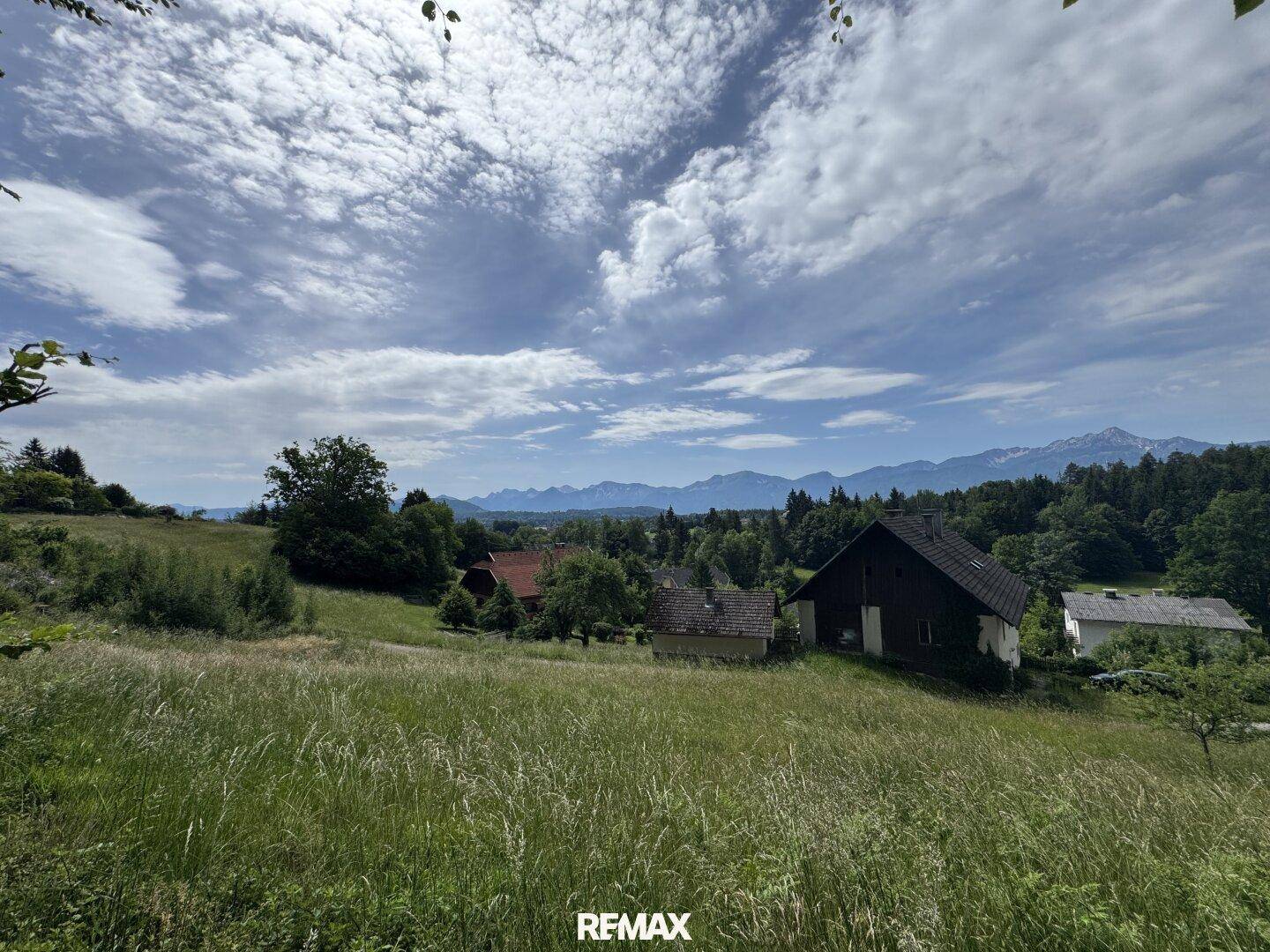 Panoramablick über eine grüne Wiese mit Häusern und Bergen am Horizont unter Wolken.