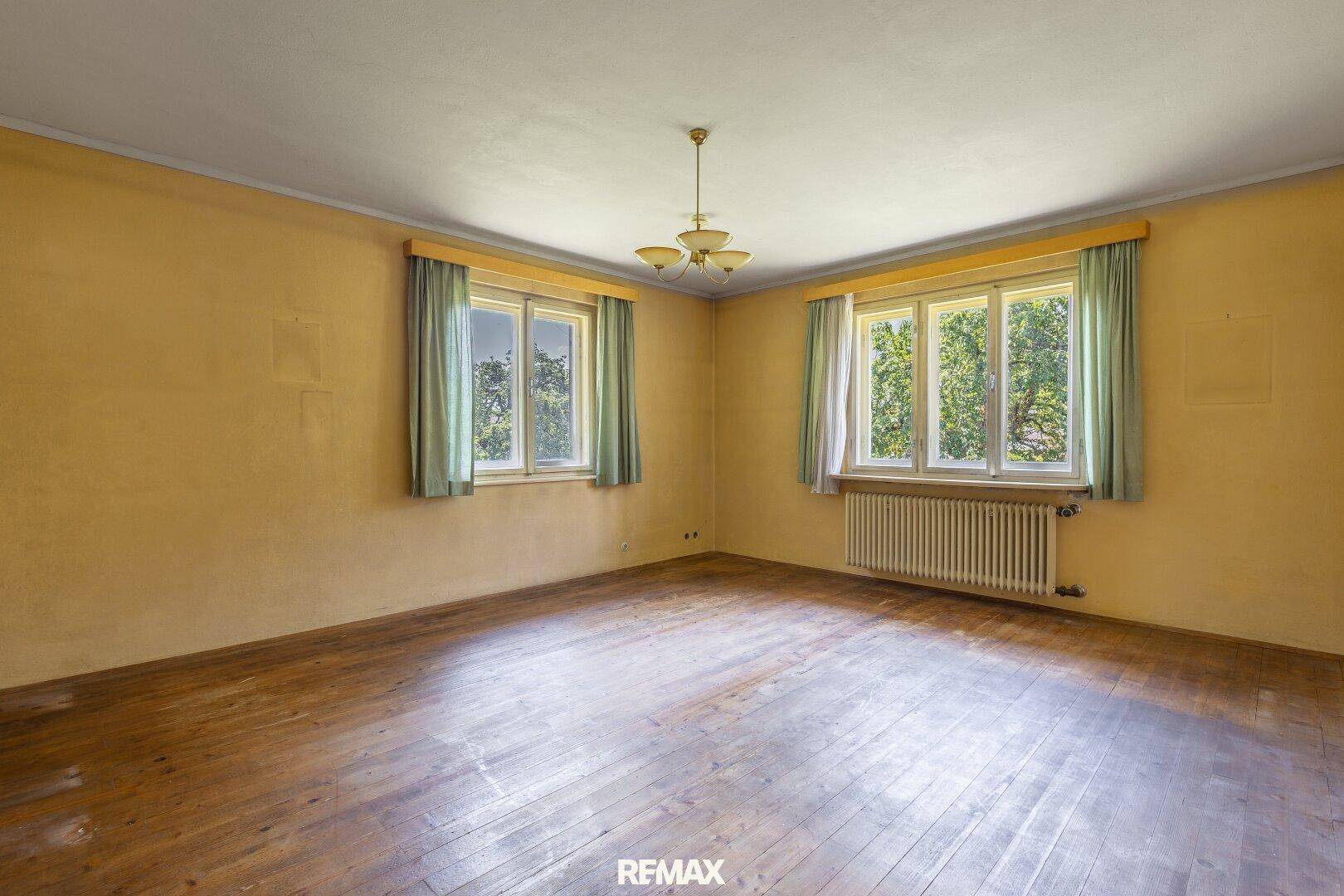 Helles Wohnzimmer mit Holzboden, zwei großen Fenstern und gelben Wänden.