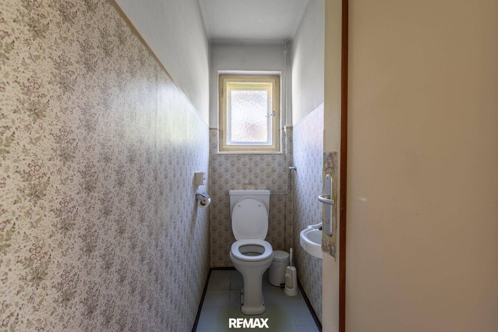 Kleines Gäste-WC mit Toilette, Waschbecken und Fenster in schmalem Raum.