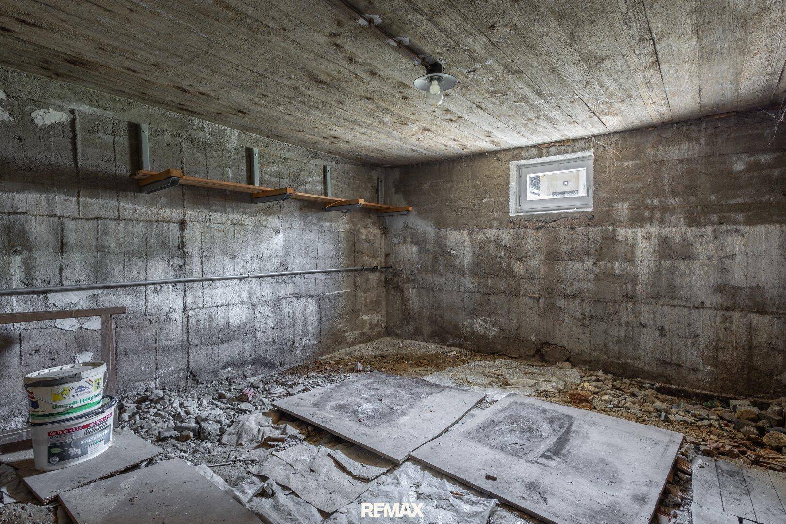Unfertiger Kellerraum mit Betonwänden, kleinem Fenster und Schutt auf dem Boden.