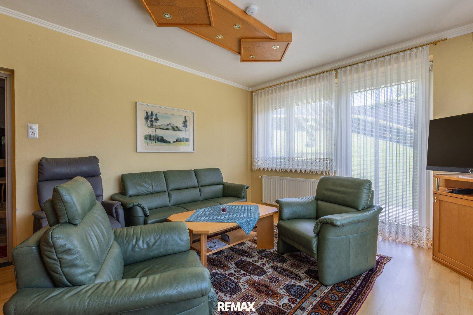 Geräumiges Wohnzimmer mit grünen Ledersofas, Holzboden und großem Fenster mit Vorhängen.