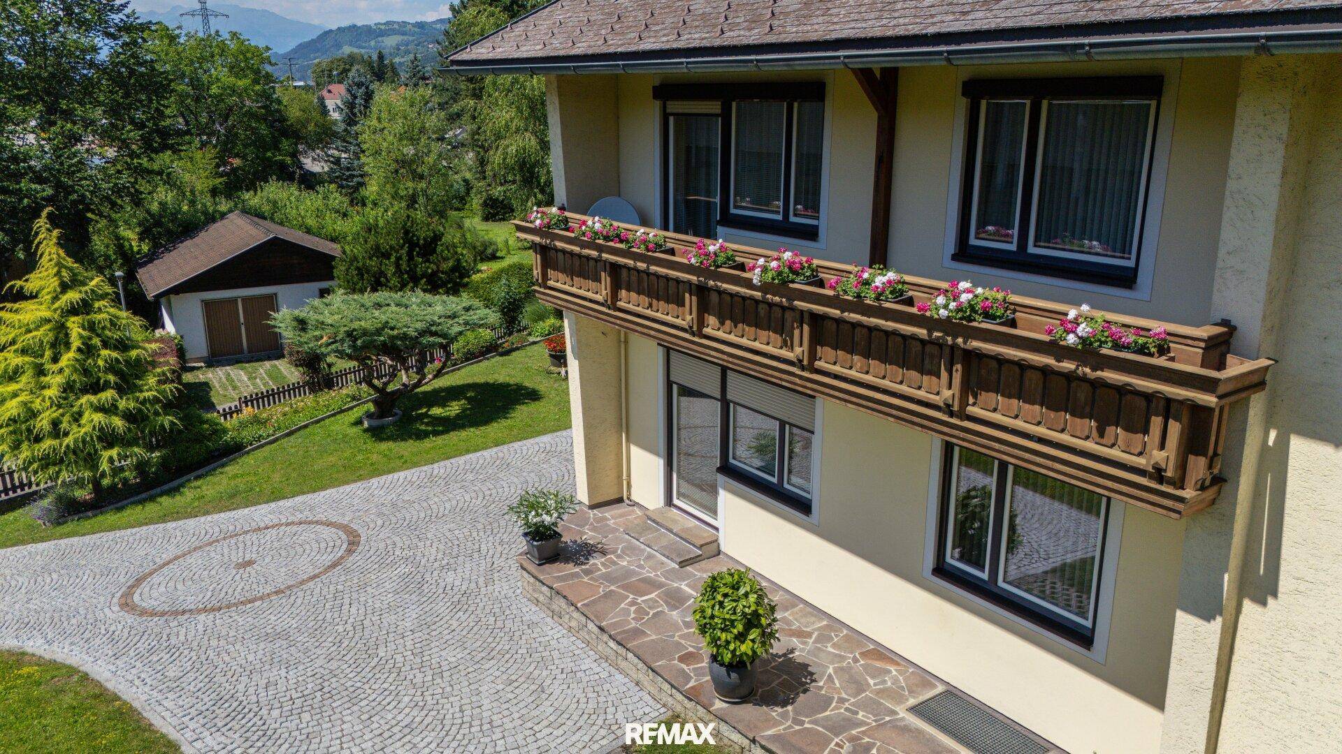 Hausfassade mit traditionellem Holzbalkon, Blumenkästen und gepflasterter Auffahrt, umgeben von Grün.