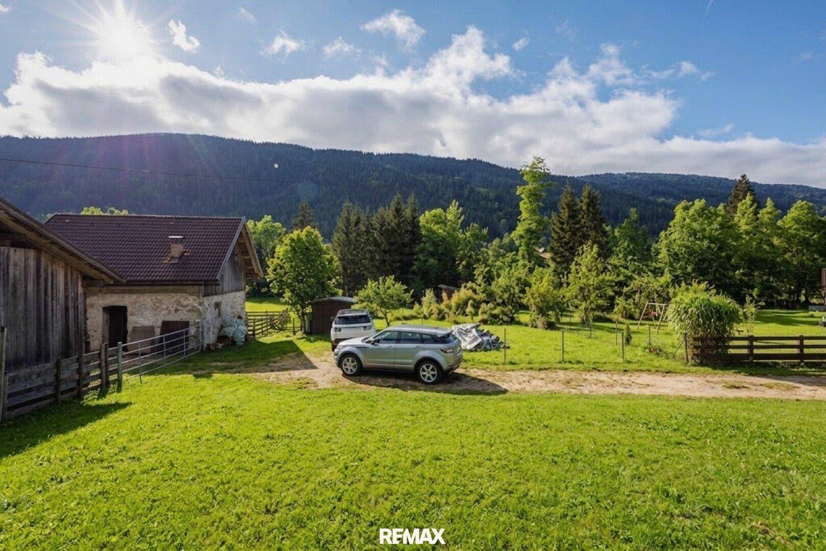Weitläufiges grünes Grundstück mit Bäumen und Bergen im Hintergrund unter blauem Himmel.