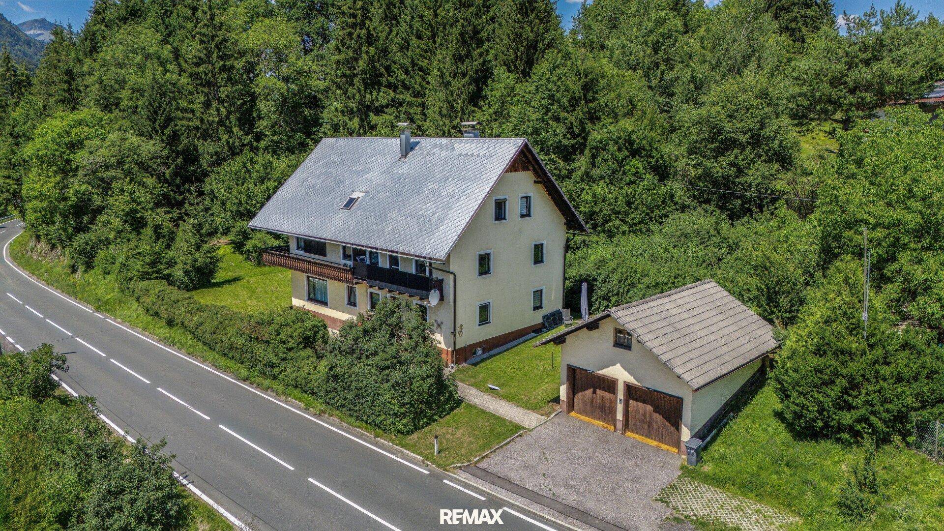 Luftaufnahme eines Hauses mit Garage, umgeben von Bäumen und einer Straße, in ländlicher Umgebung.