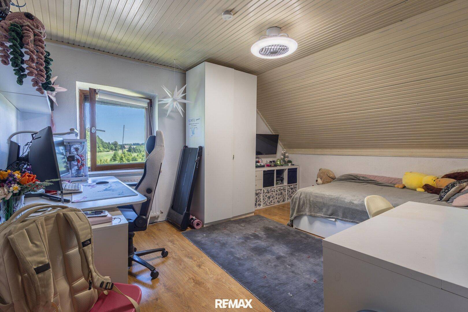 Helles Dachgeschosszimmer mit Schreibtisch, Bett und schräger Decke, bietet einen grünen Ausblick.