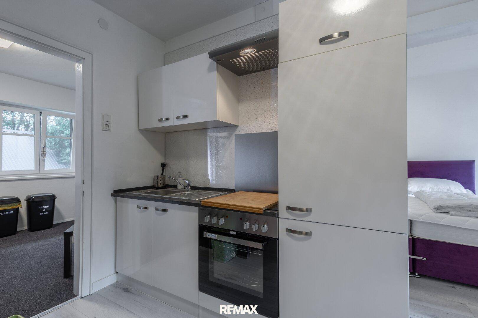 Kompakte Küchenzeile mit weißen Schränken, integriertem Backofen und Spüle, angrenzend an ein Schlafzimmer.