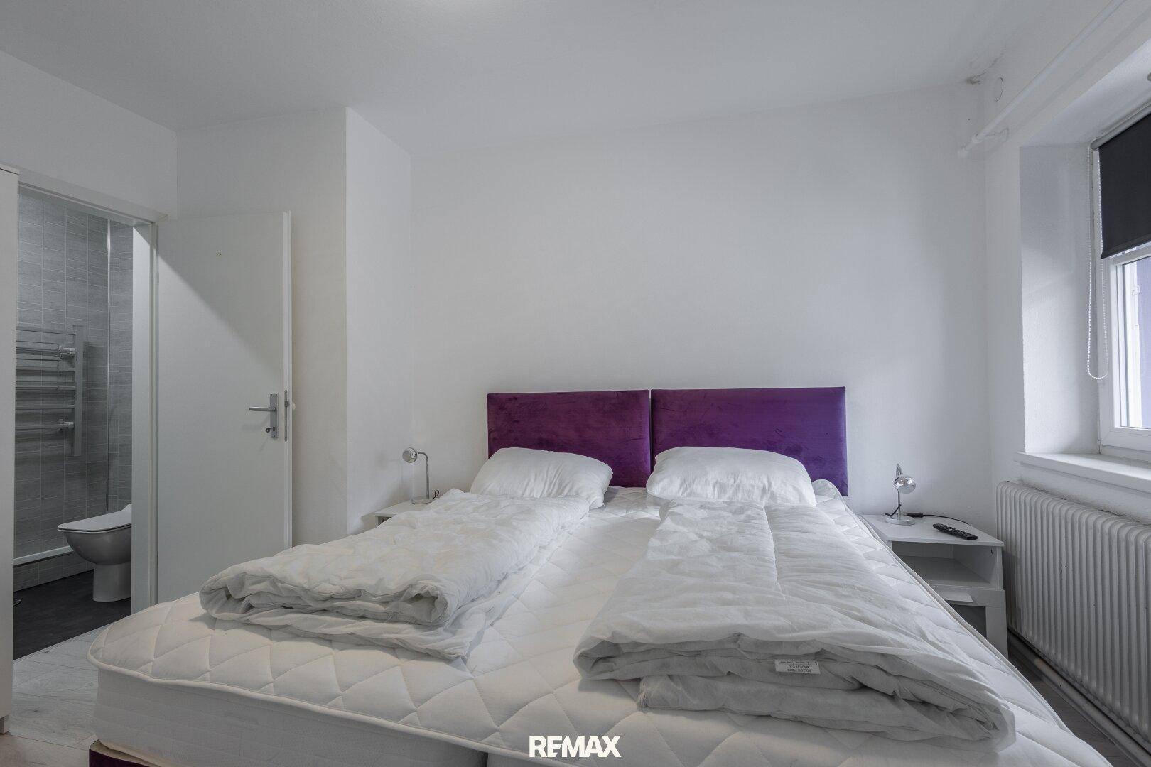 Schlafzimmer mit Doppelbett und lila Kopfteil, direktem Zugang zu einem modernen Badezimmer.