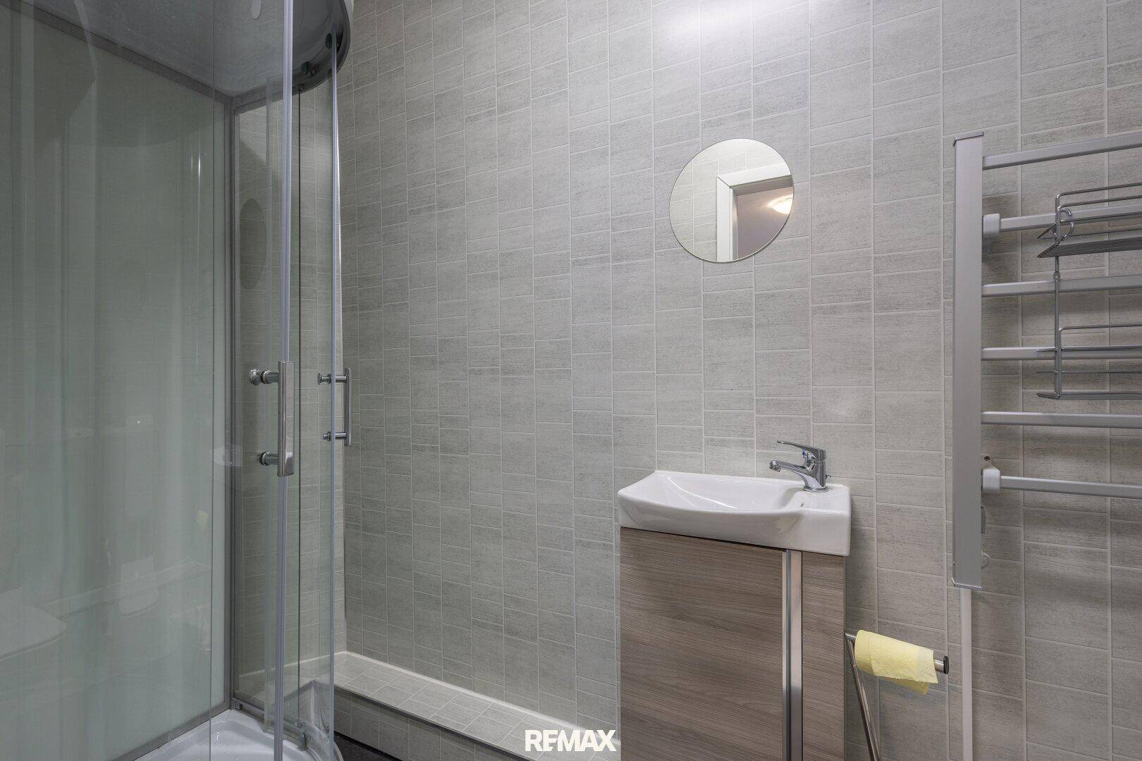 Modernes Badezimmer mit grauen Fliesen, Duschkabine, Einzelwaschbecken und beheiztem Handtuchhalter.