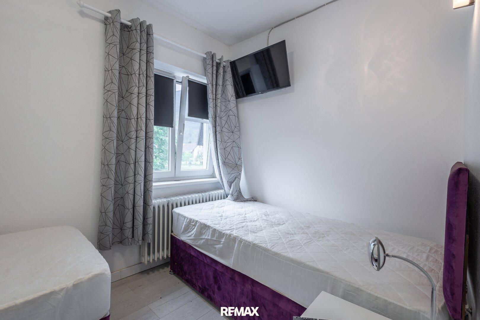 Helles Schlafzimmer mit Einzelbett, Fenster und Fernseher an der Wand.