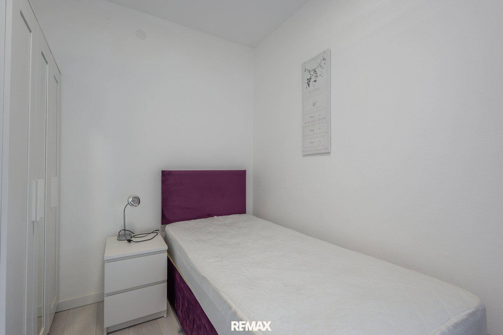 Schlafzimmer mit Einzelbett, lila Kopfteil und weißem Kleiderschrank.