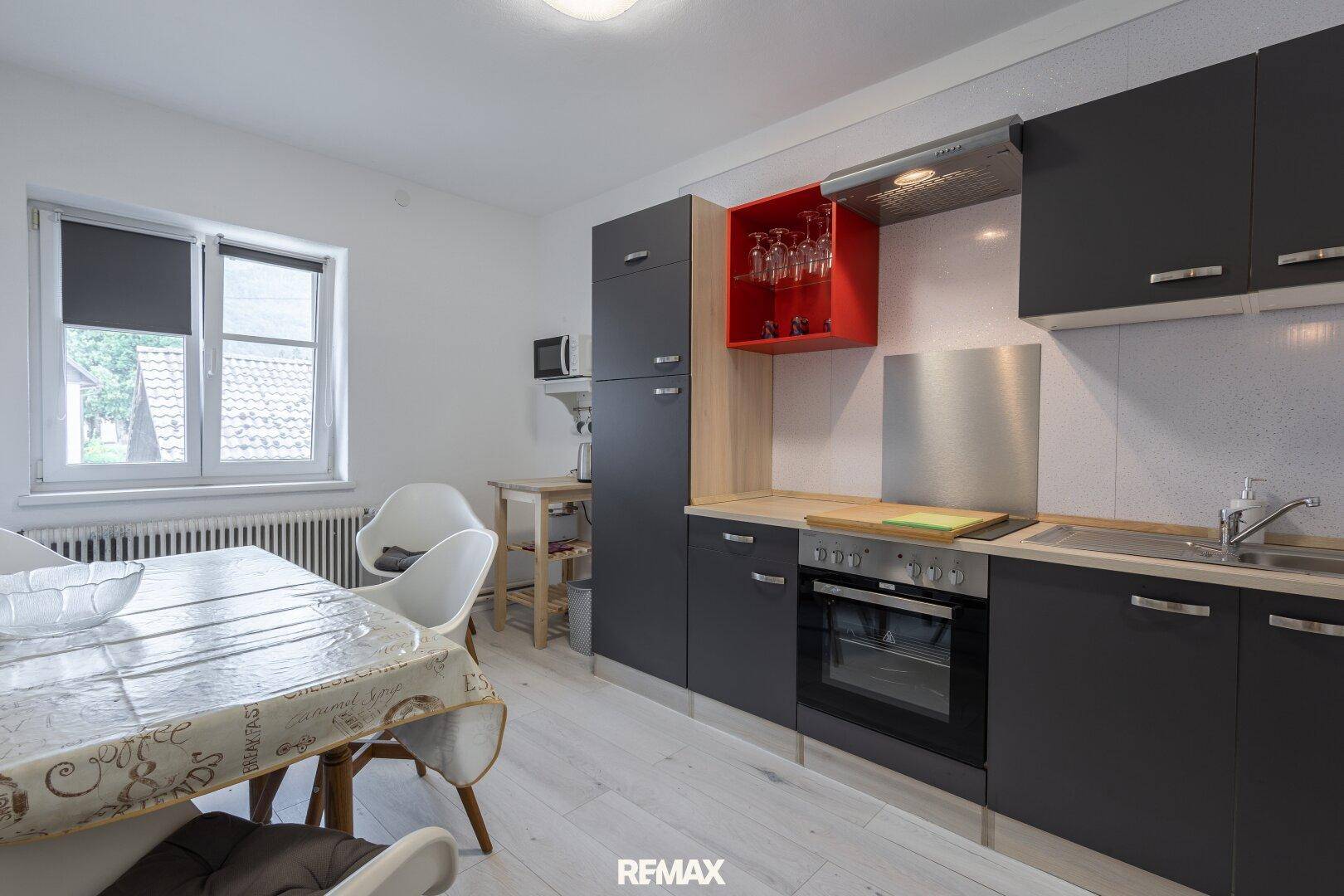 Moderne Küche mit grauen Schränken, Essbereich und Fenster, ausgestattet mit Backofen und Dunstabzugshaube.
