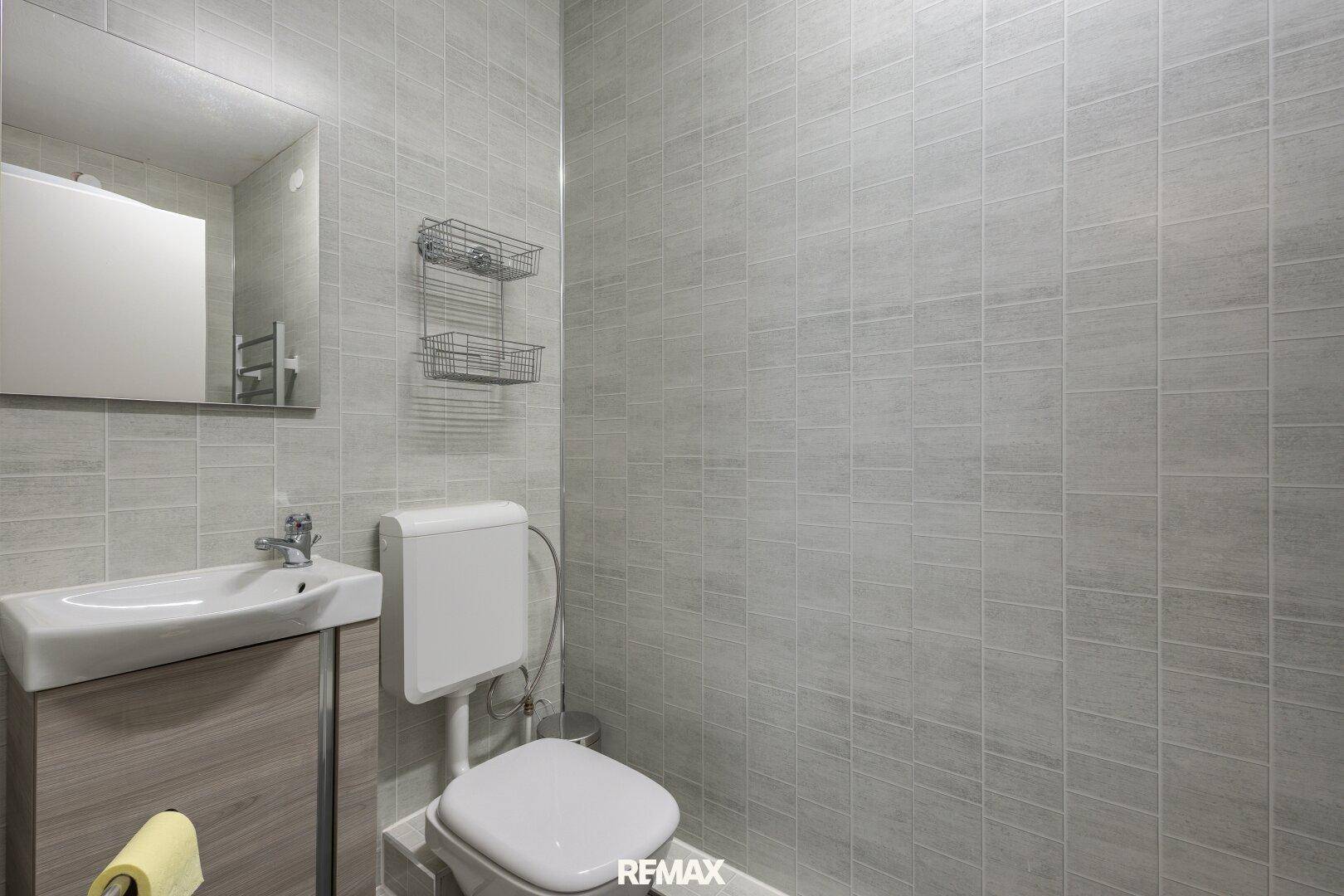 Kleines Badezimmer mit Toilette, Einzelwaschbecken und Spiegel, komplett gefliest in Grau.