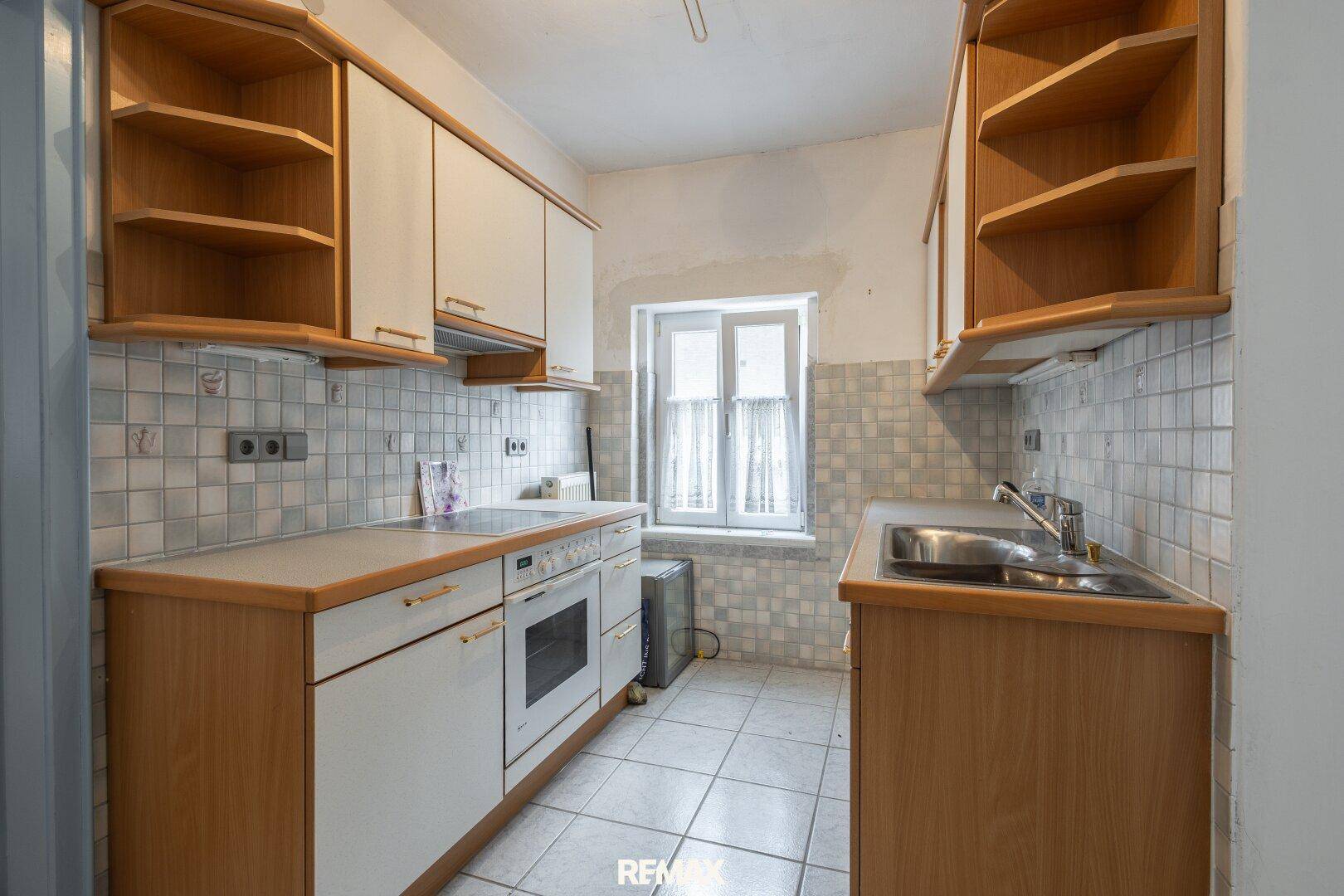 Ältere Küche mit Holzschränken, Fliesenboden und Fenster, die eine Renovierung benötigt.