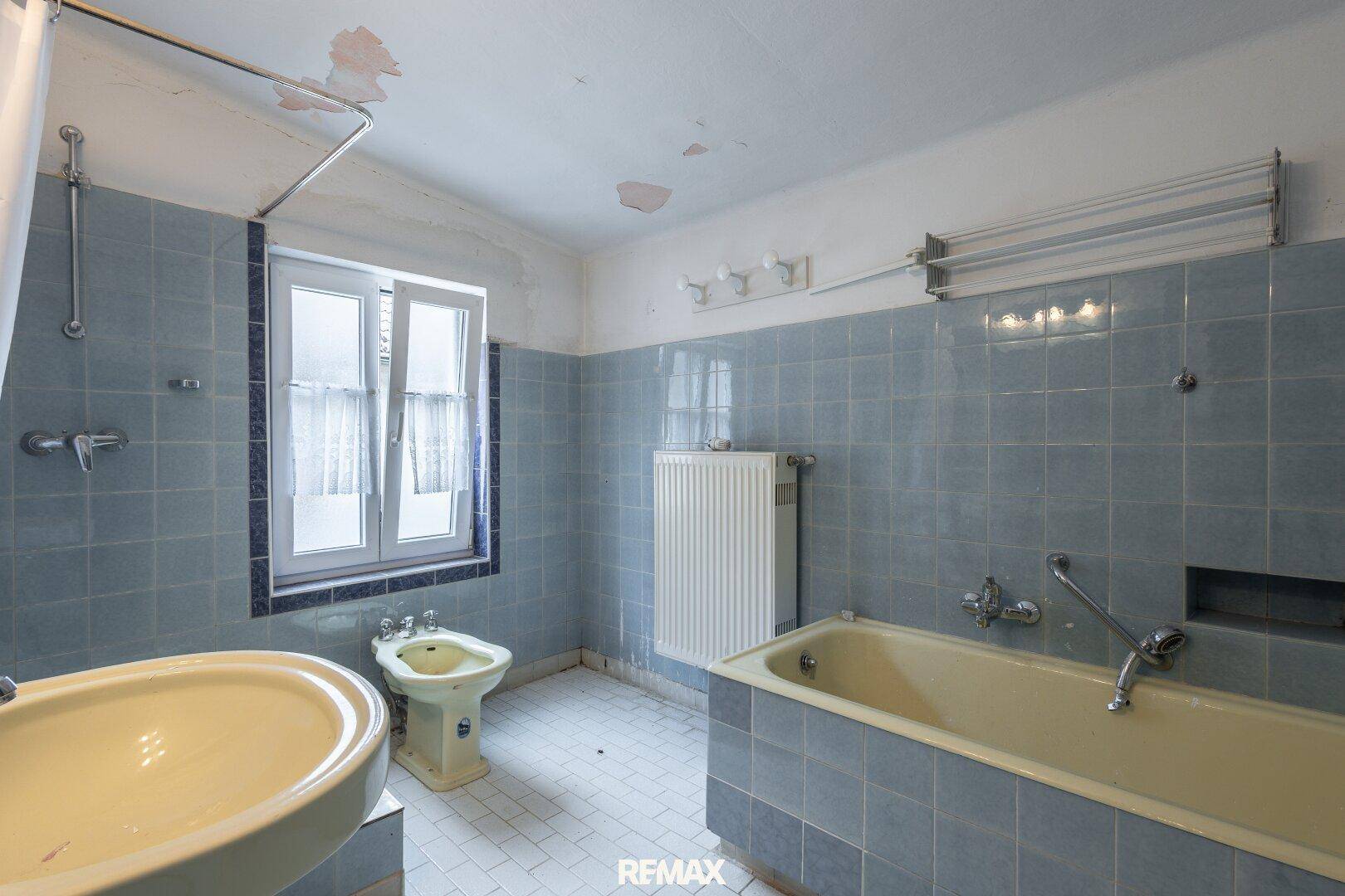 Veraltetes Badezimmer mit blauen Fliesen, Badewanne, Bidet und Fenster, das renovierungsbedürftig ist.