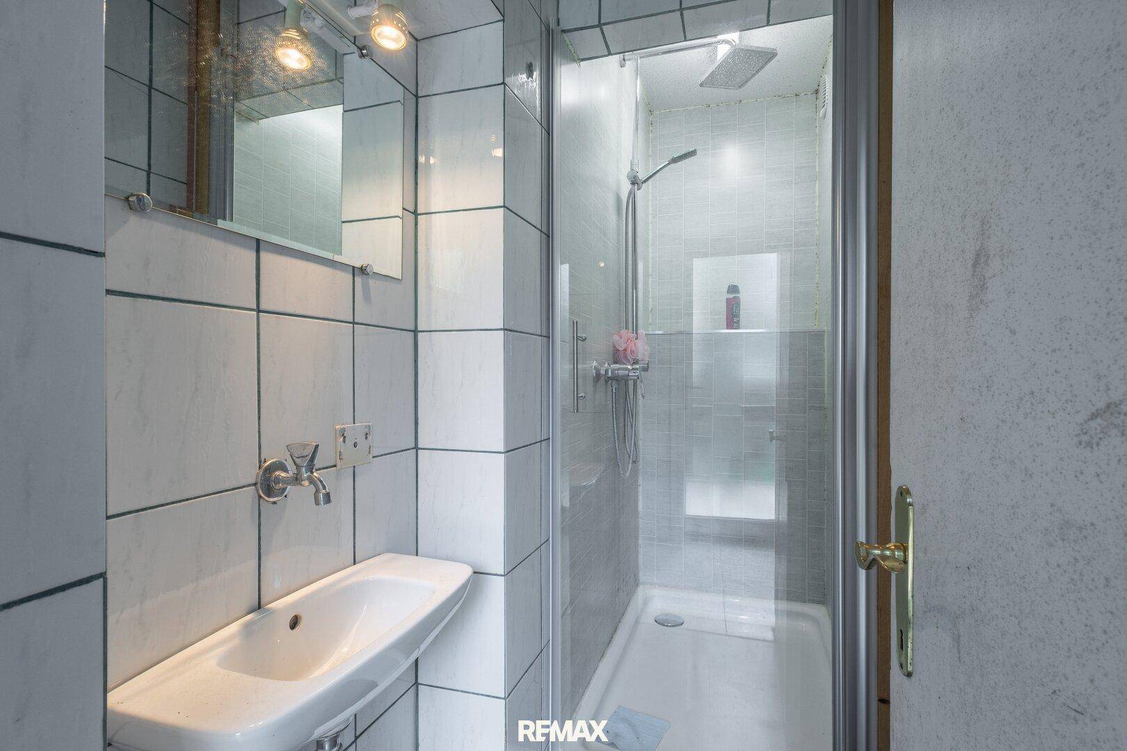 Kleines Badezimmer mit weißen Fliesen, Waschbecken und integrierter Duschkabine.