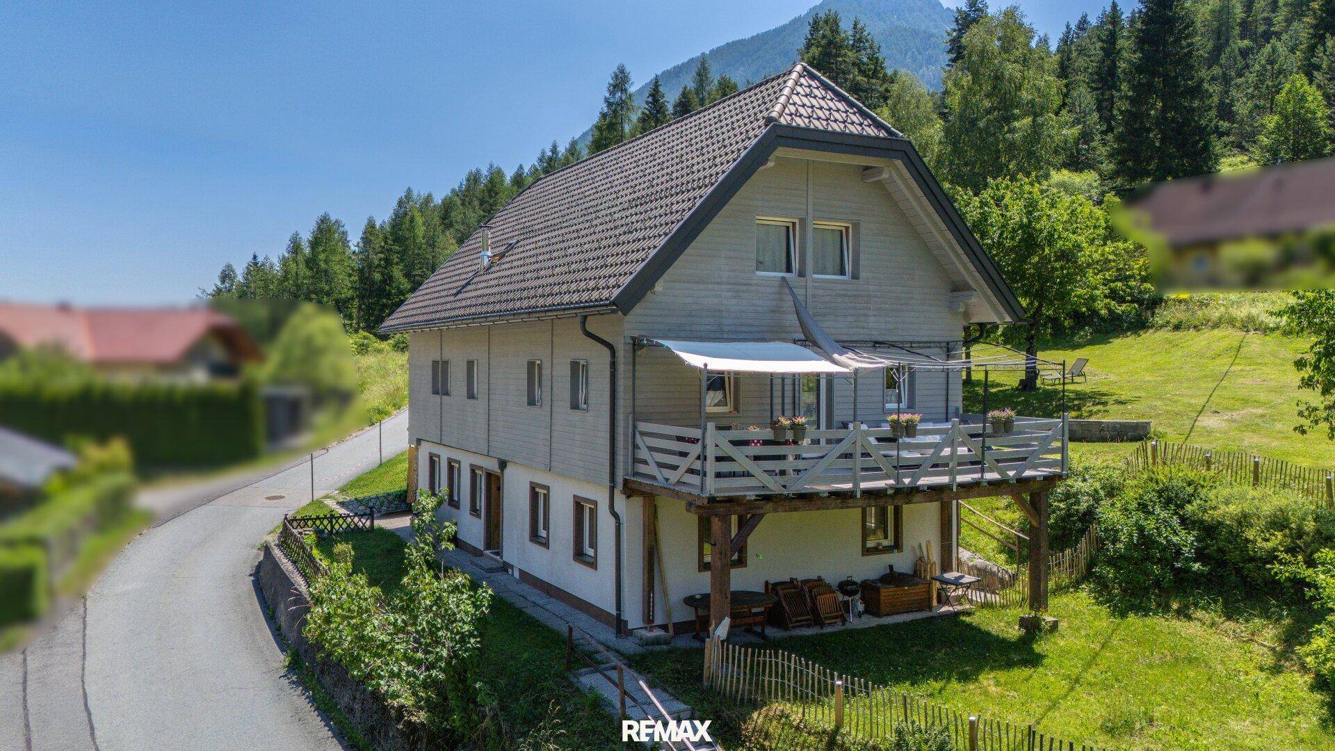 Haus mit Holzverkleidung und Balkon, umgeben von grüner Natur und mit Bergblick.
