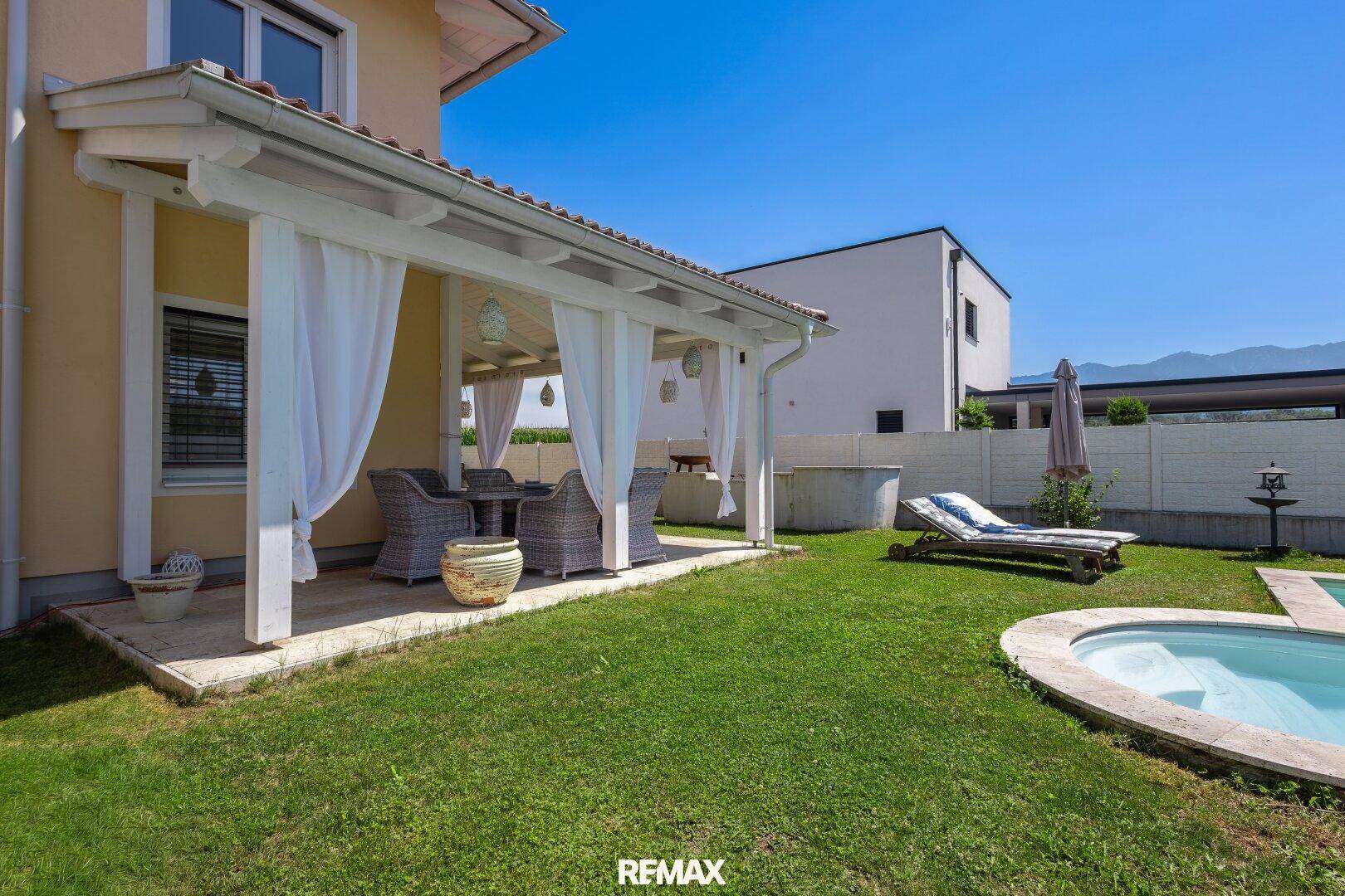 Überdachte Terrasse mit Rattanmöbeln und Blick auf den Poolbereich im sonnigen Garten.