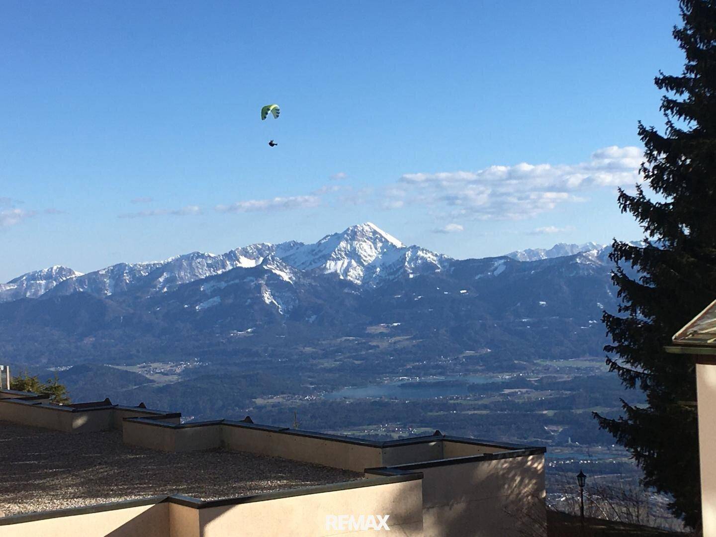 Panoramablick auf schneebedeckte Berge und ein Tal mit See unter blauem Himmel.