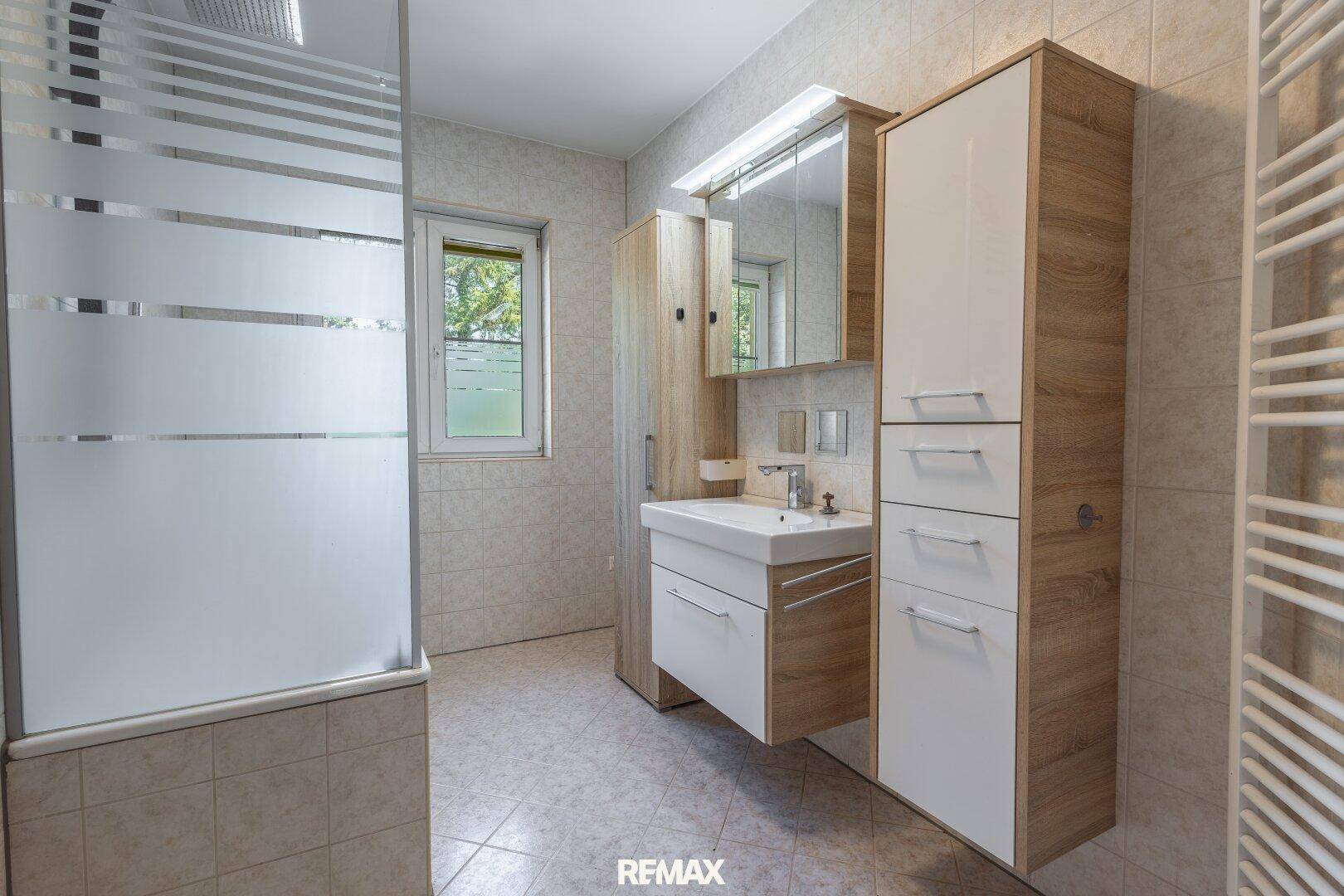 Modernes Badezimmer mit Dusche, Waschbecken und Spiegelschrank, hell gefliest und mit Fenster.