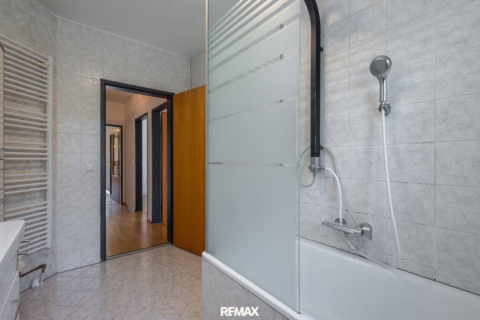 Badezimmer mit Badewanne und Duschvorrichtung, hellen Fliesen und praktischem Handtuchheizkörper.