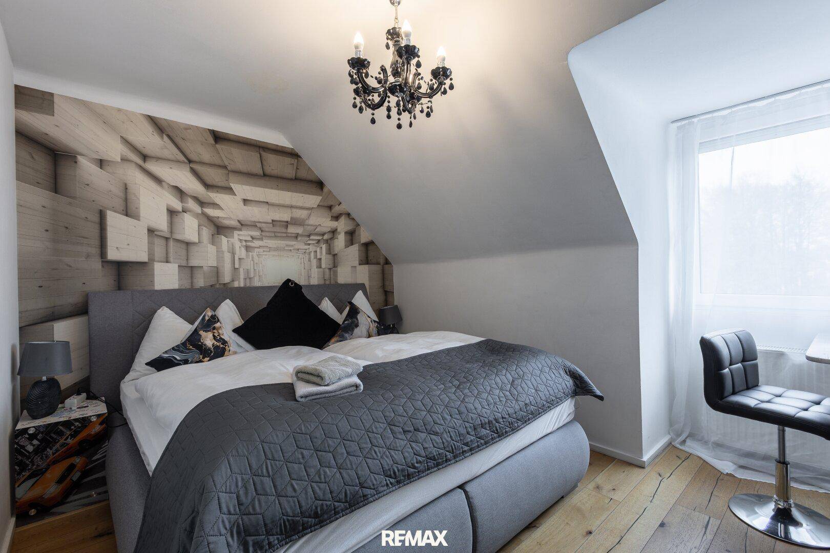 Dachgeschoss-Schlafzimmer mit schräger Decke, Doppelbett und moderner geometrischer Tapete.