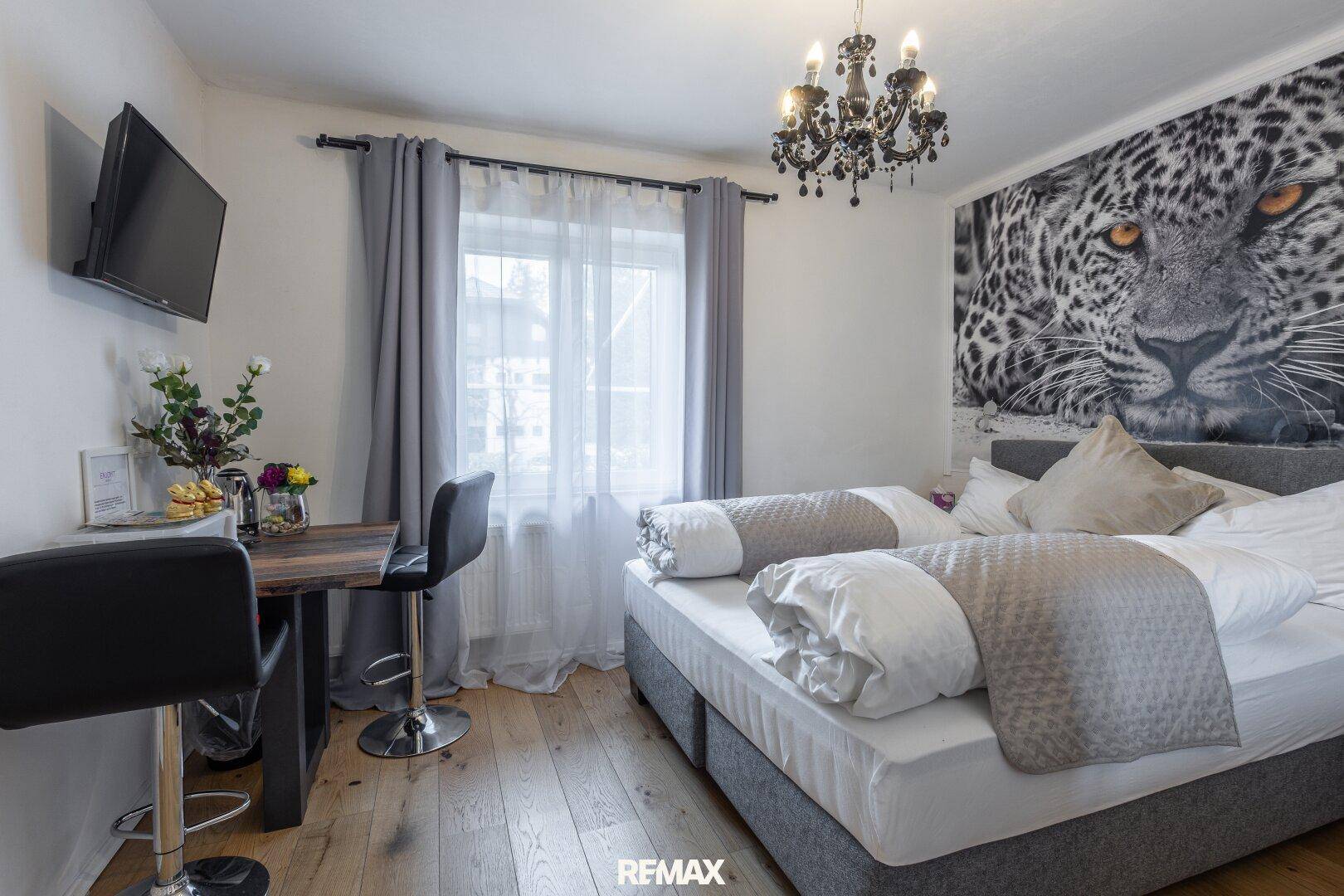 Schlafzimmer mit Doppelbett, Leopard-Wandbild, Holzboden und kleinem Tisch mit Barhockern.