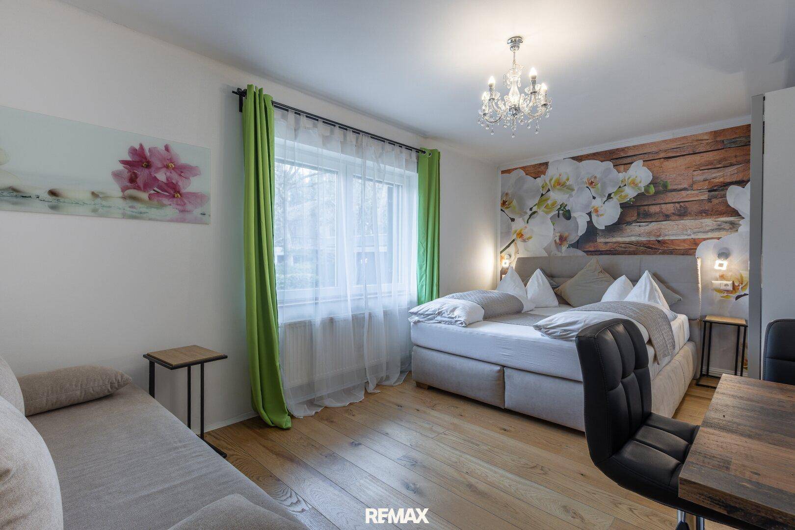 Helles Schlafzimmer mit Doppelbett, floraler Wanddekoration, Holzboden und grünen Vorhängen.