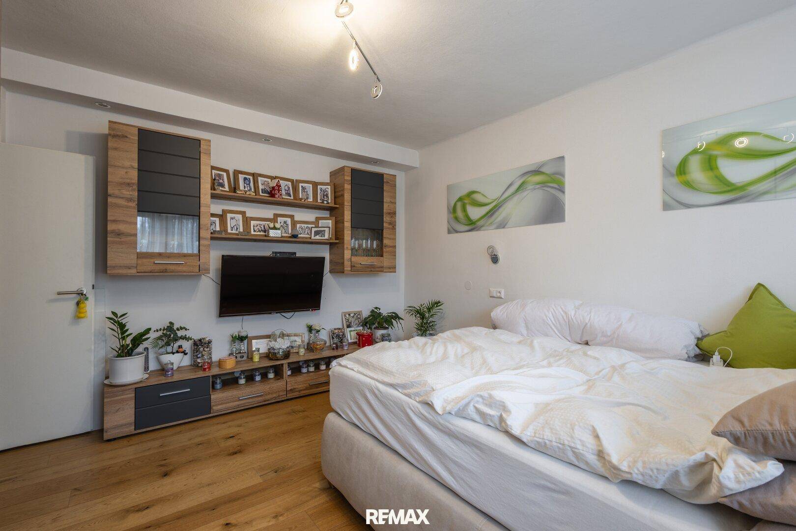 Schlafzimmer mit Doppelbett, Holzboden und einer modernen TV-Wand mit Regalen und Dekoration.
