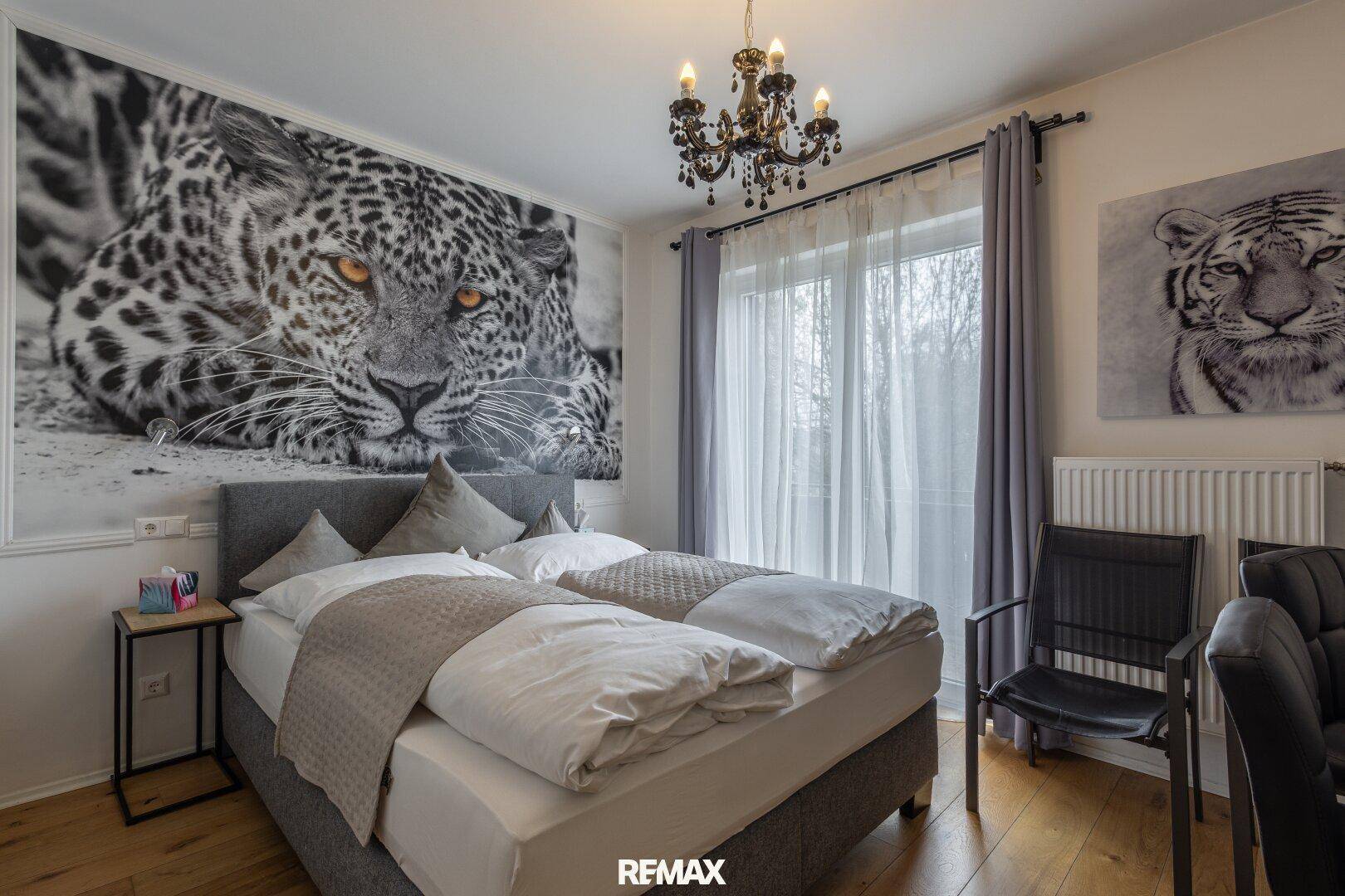 Helles Schlafzimmer mit Doppelbett, Leopard-Wandbild und einem kleinen Tisch mit Stuhl.