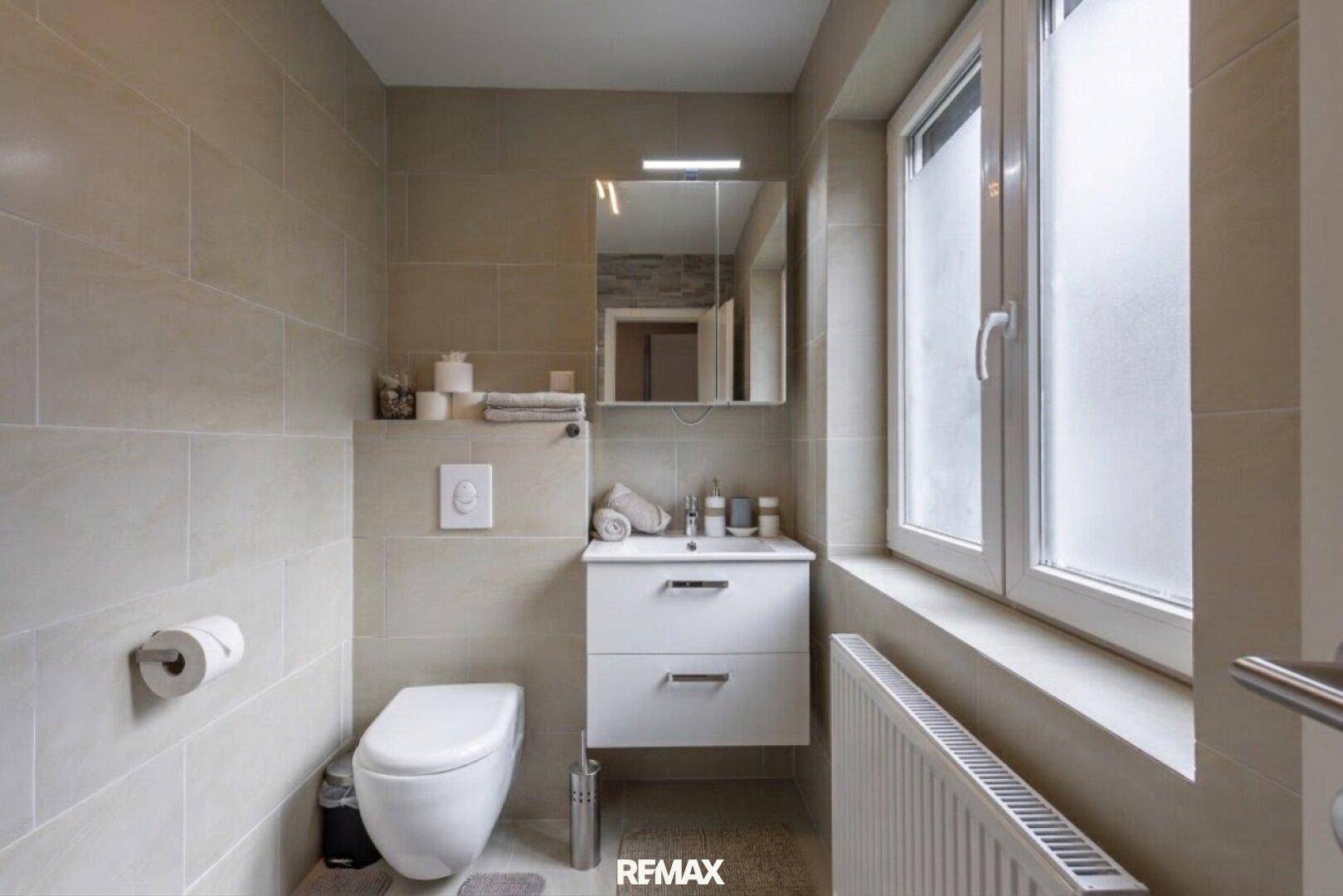 Helles Badezimmer mit Toilette, Waschbecken, Spiegelschrank und Fenster für Tageslicht.
