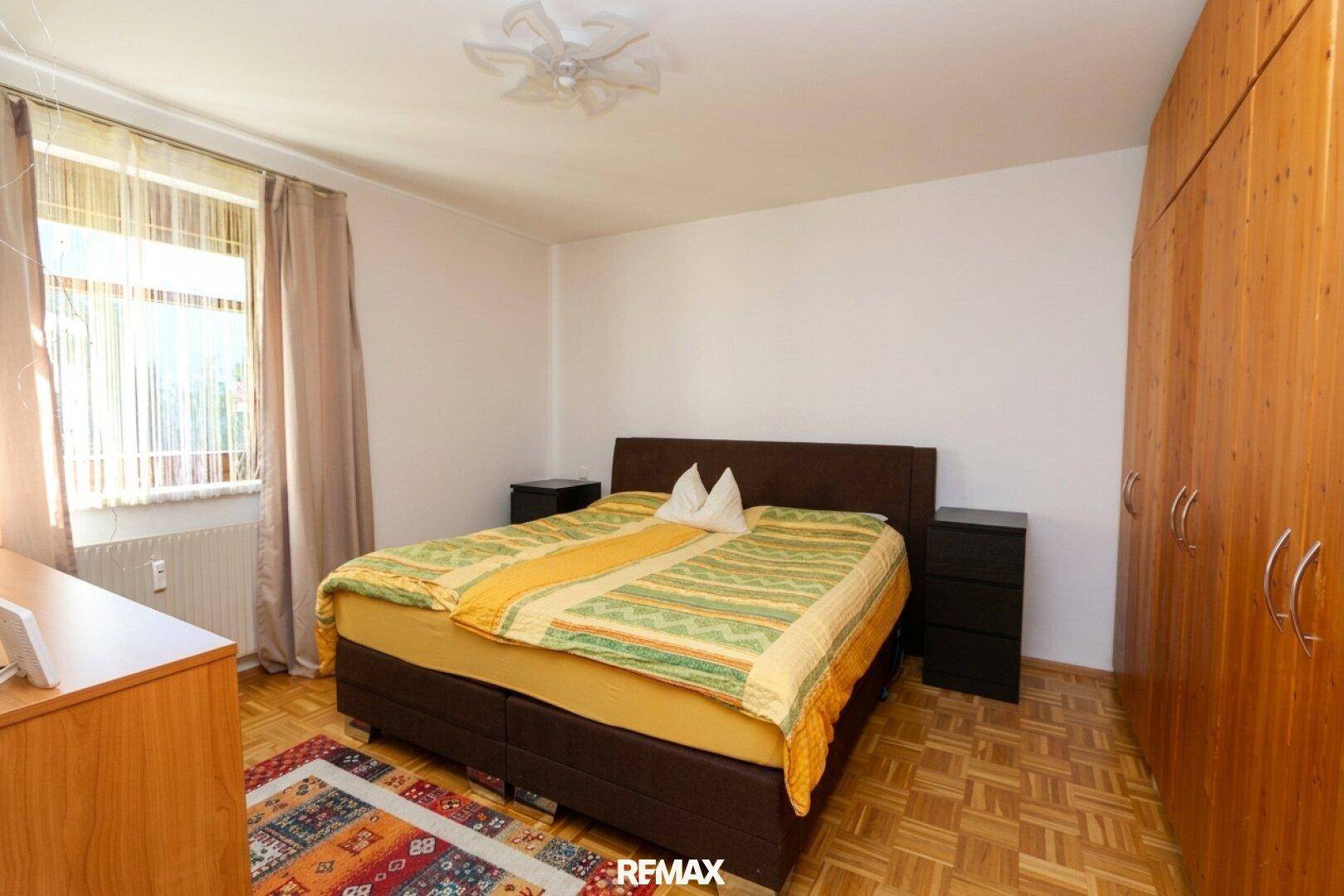Schlafzimmer mit Doppelbett, Holzschrank und Parkettboden, gut beleuchtet durch ein Fenster.