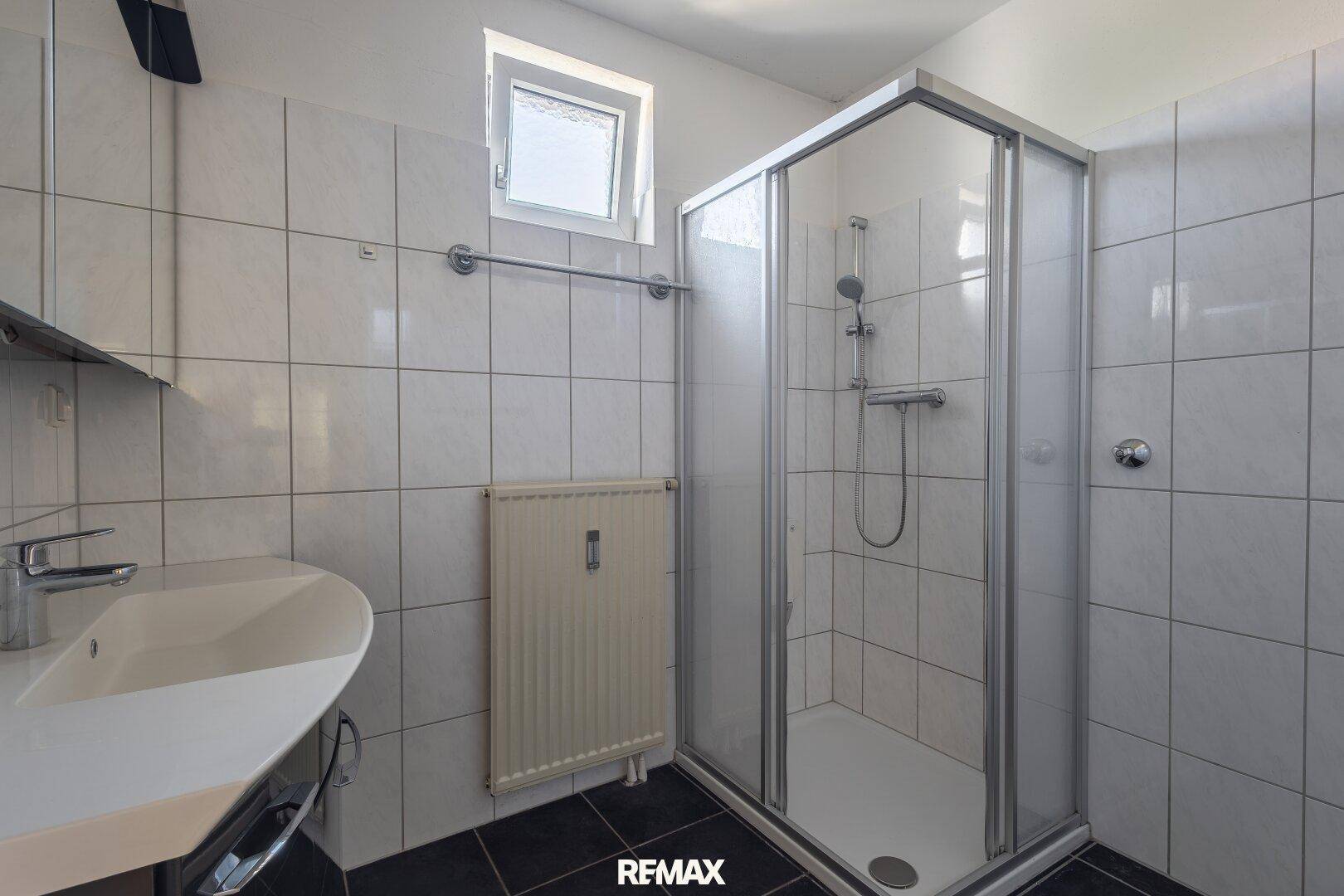 Helles Badezimmer mit Duschkabine, Waschbecken und weißen Fliesen, ausgestattet mit einem Fenster.