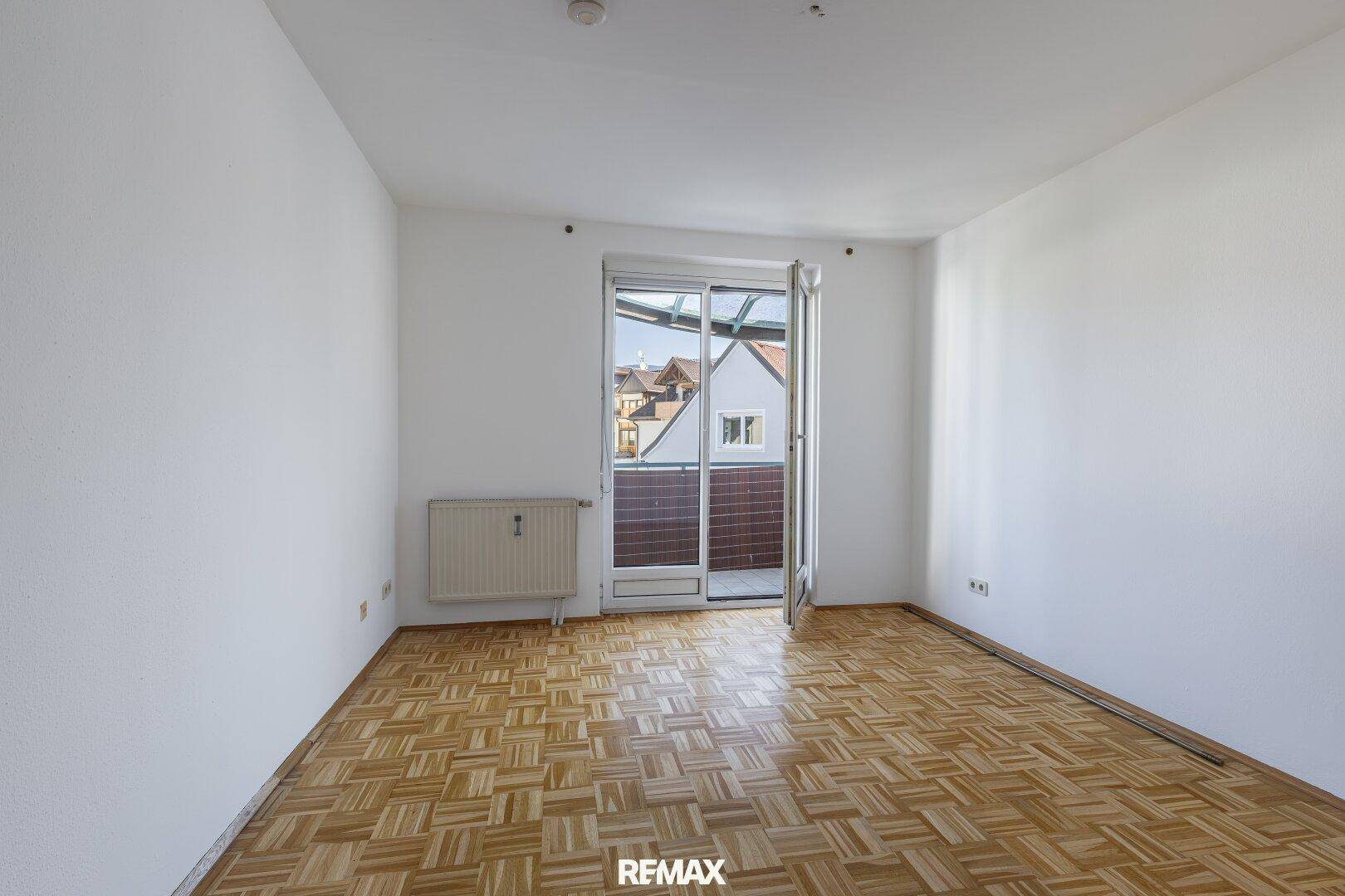Leeres Schlafzimmer mit Parkettboden und Balkonzugang, das viel natürliches Licht hereinlässt.