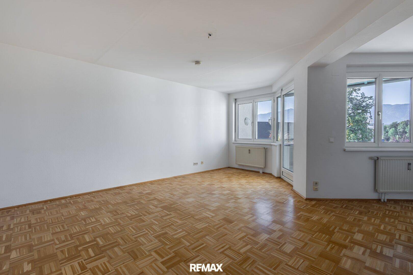 Leeres Wohnzimmer mit Parkettboden und großen Fenstern, die einen Blick auf die Berge bieten.