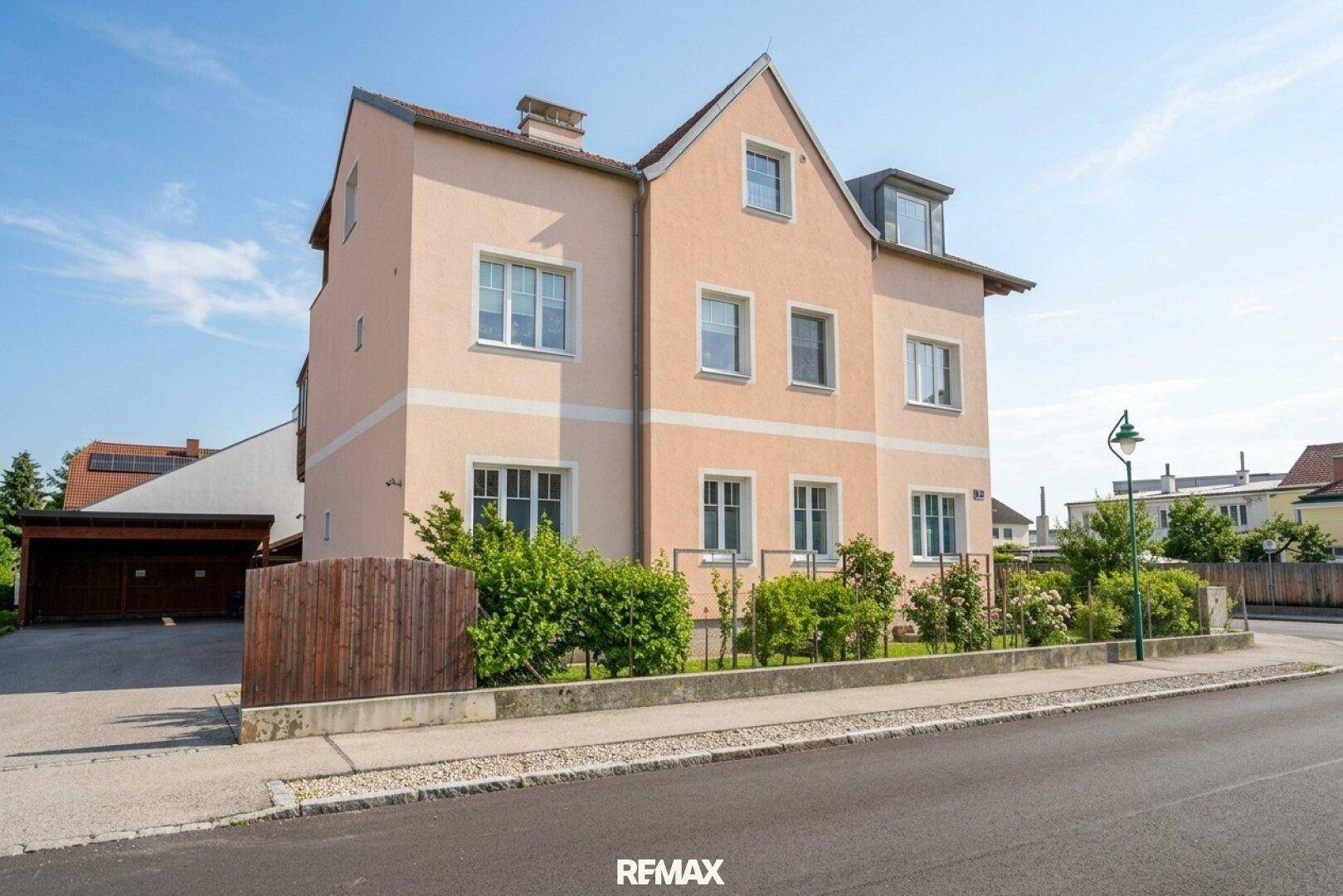 Ansicht des Gebäudes von der Straßenseite mit hellrosa Fassade und kleinen Büschen im Vorgarten.