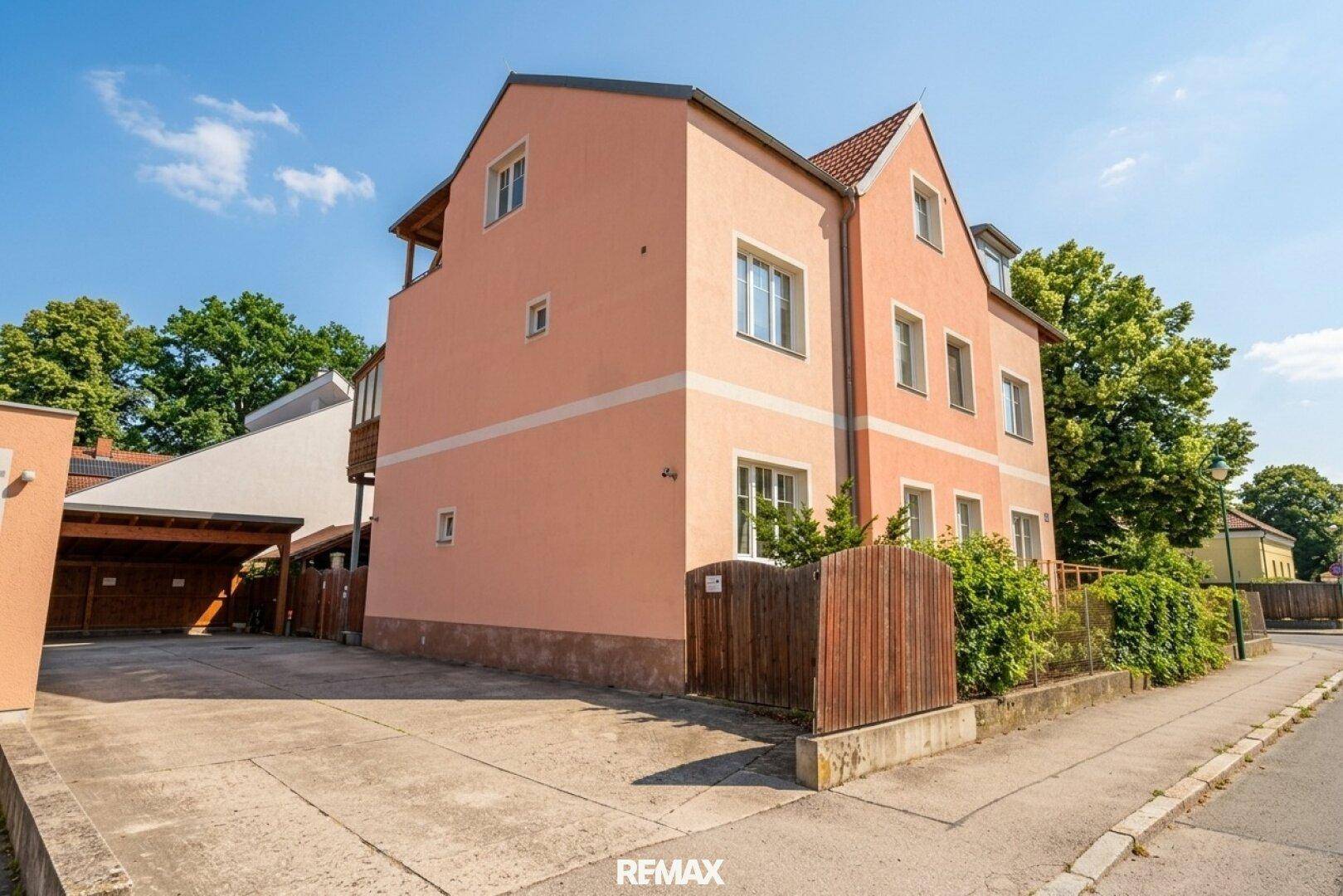 Seitenansicht des Gebäudes mit hellrosa Fassade, einem Balkon und einer Zufahrt zum Parkbereich.