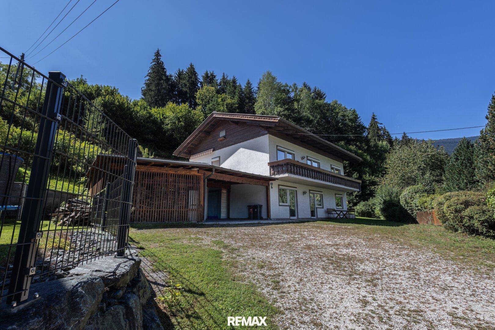 Gesamtansicht des Hauses mit Carport, großem Vorplatz und umzäuntem Grundstück in grüner Umgebung.