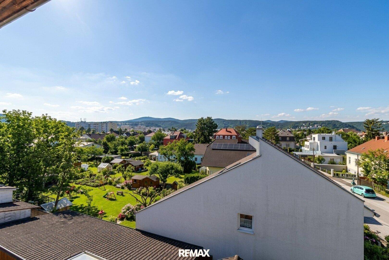 Weitläufige Aussicht über Gärten und Häuser, mit grünen Hügeln im Hintergrund unter blauem Himmel.
