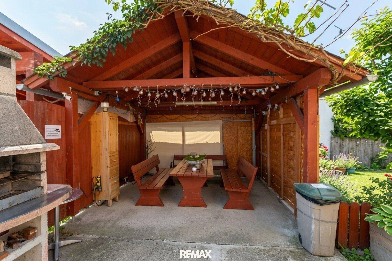 Überdachter Grillplatz mit Holztisch und Bänken, umgeben von Grün und mit Lichterketten dekoriert.