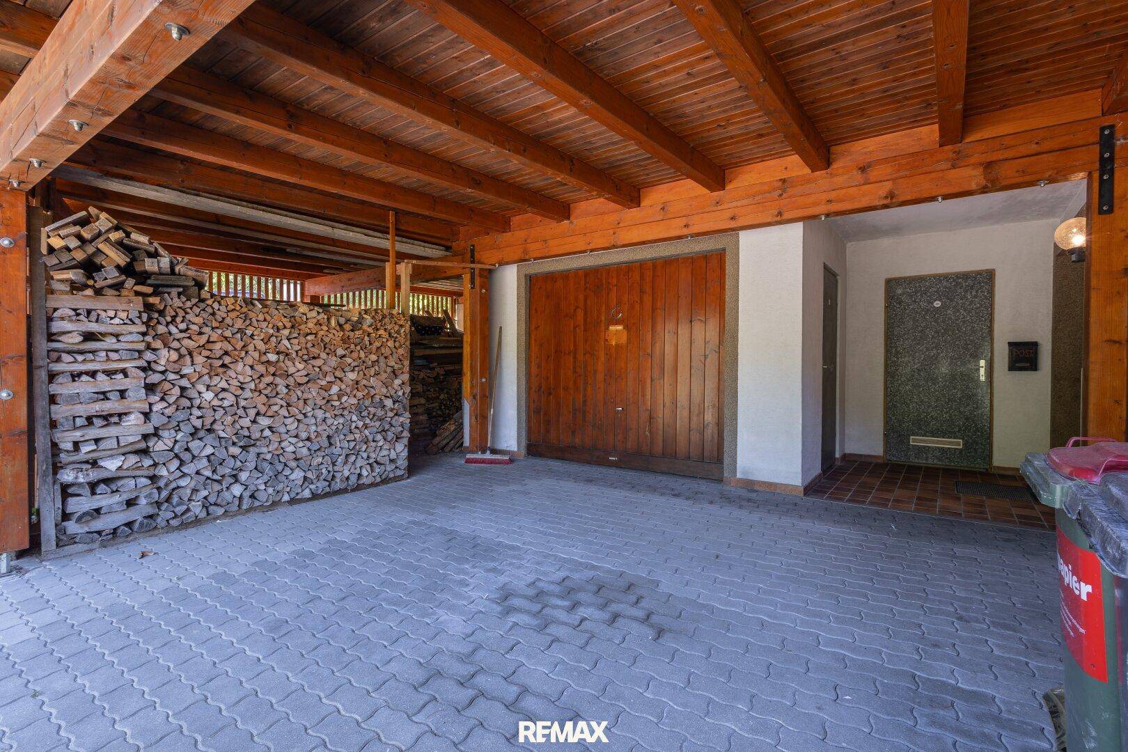 Überdachter Carport mit Holzbalken und Lagerplatz für Brennholz neben einer Garage.