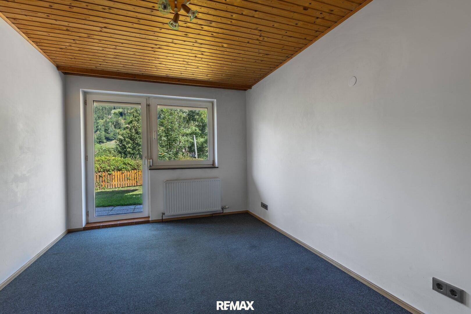 Schlafzimmer mit blauem Teppichboden, Holzdecke und Zugang zu einem kleinen Gartenbereich.