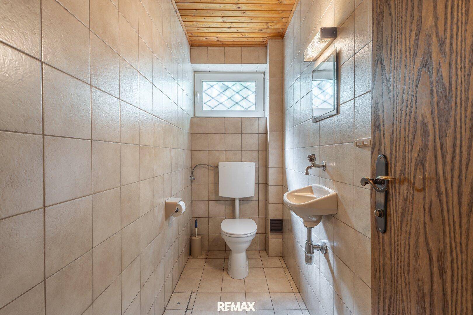 Kleines WC mit Fliesenboden, Waschbecken und Fenster, einfach und funktional.