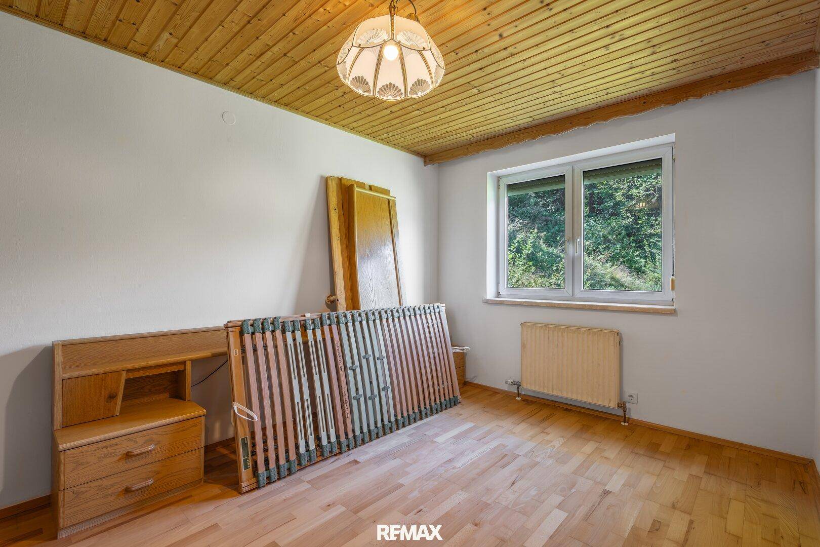 Leeres Schlafzimmer mit Holzboden, Holzdecke und Fenster mit Blick ins Grüne.