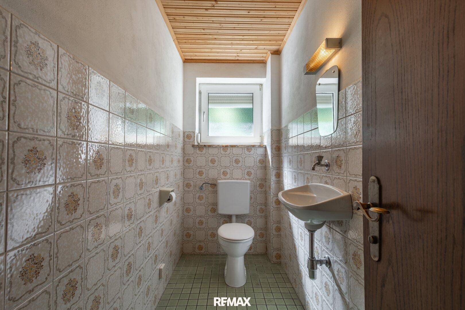 Kleines Badezimmer mit WC, Waschbecken, gemusterten Fliesen und Holzdecke in älterem Stil.