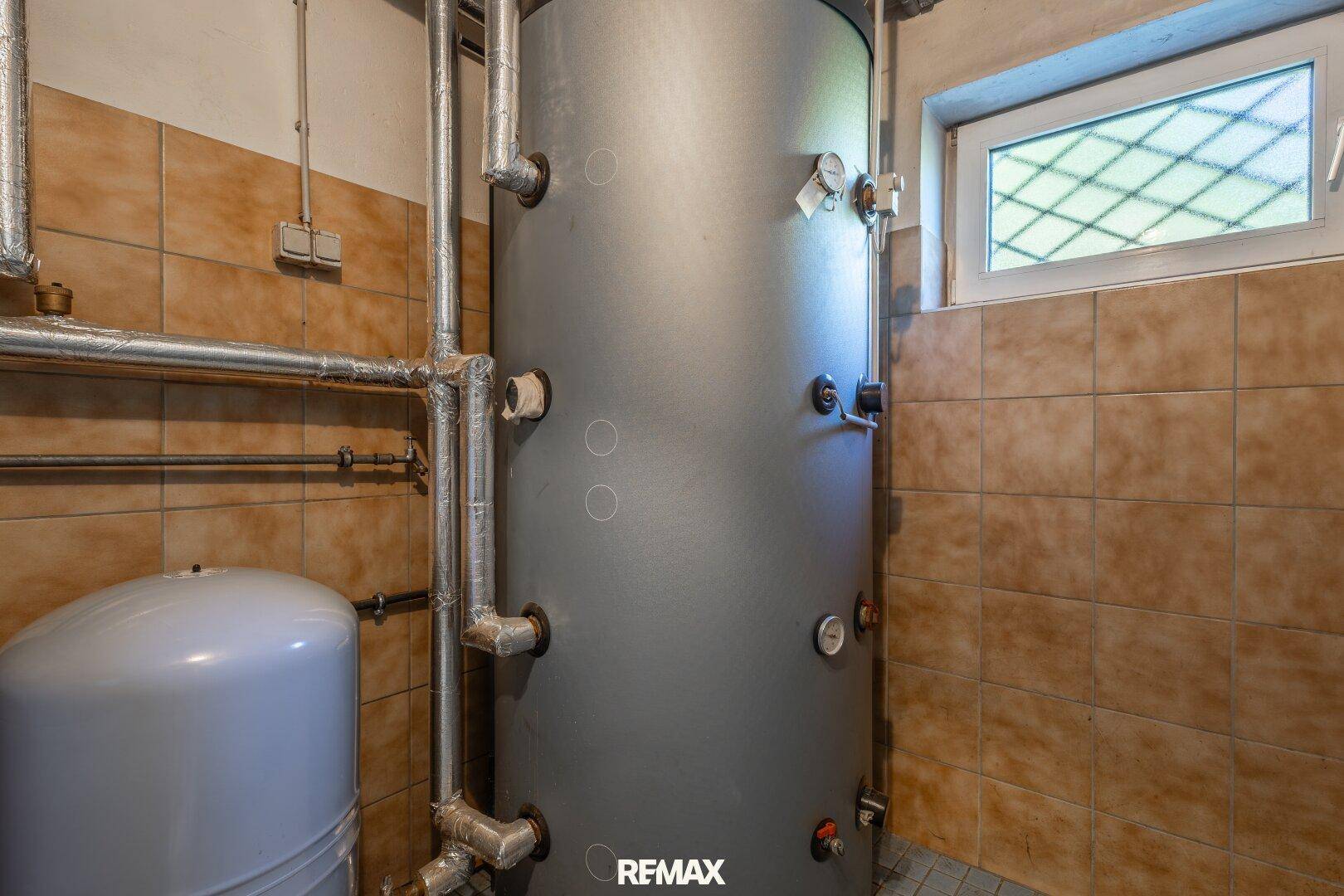 Großer Warmwasserspeicher im Heizungsraum mit Anschlüssen und einem kleinen Fenster.