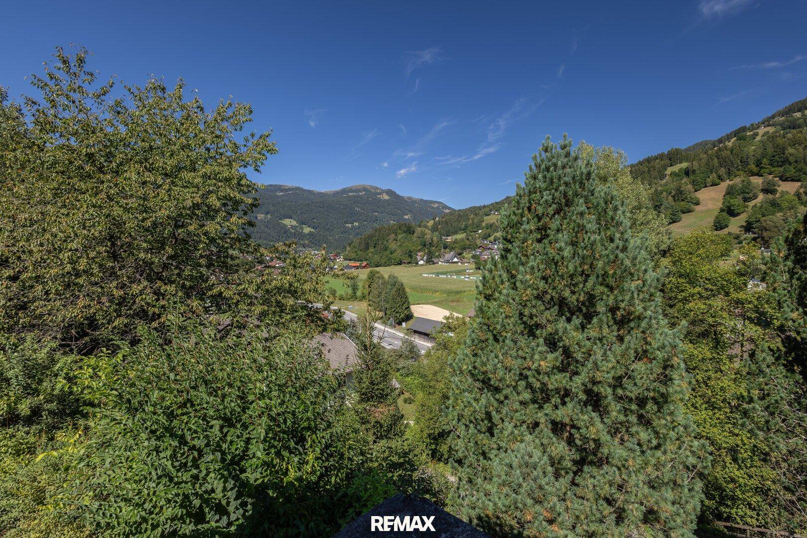 Malerischer Ausblick auf grüne Berge und ein Tal mit vereinzelten Häusern unter blauem Himmel.