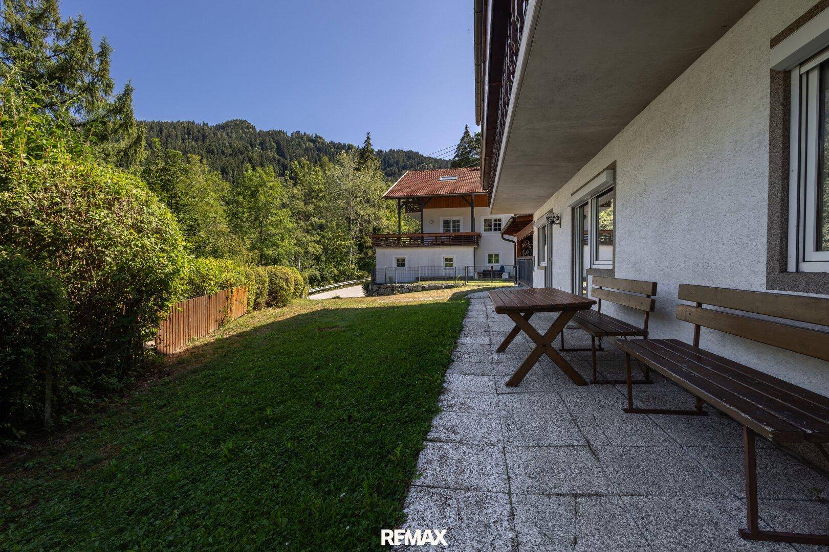 Gemütliche Terrasse mit Holzbänken und Tisch, angrenzend an einen grünen Rasenbereich.